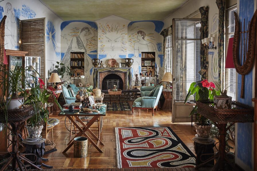 A living room with artistic wall murals, wicker furniture, plants, bookshelves, and a fireplace. A patterned rug lies on the wooden floor, and large windows bring in natural light.
