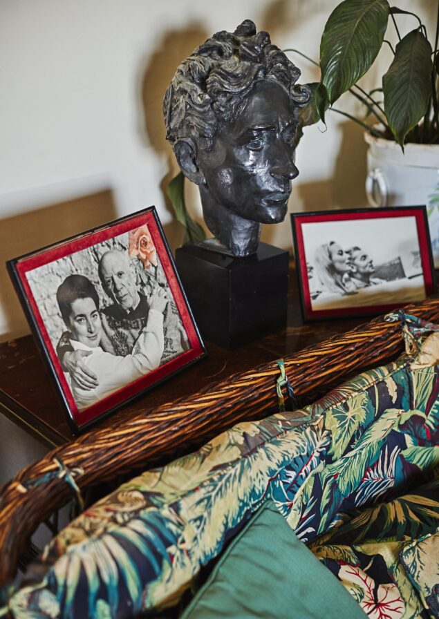 A bust sculpture is placed on a table with two framed photos beside it. A patterned cushion on a wicker chair is in the foreground, and a potted plant is in the background.