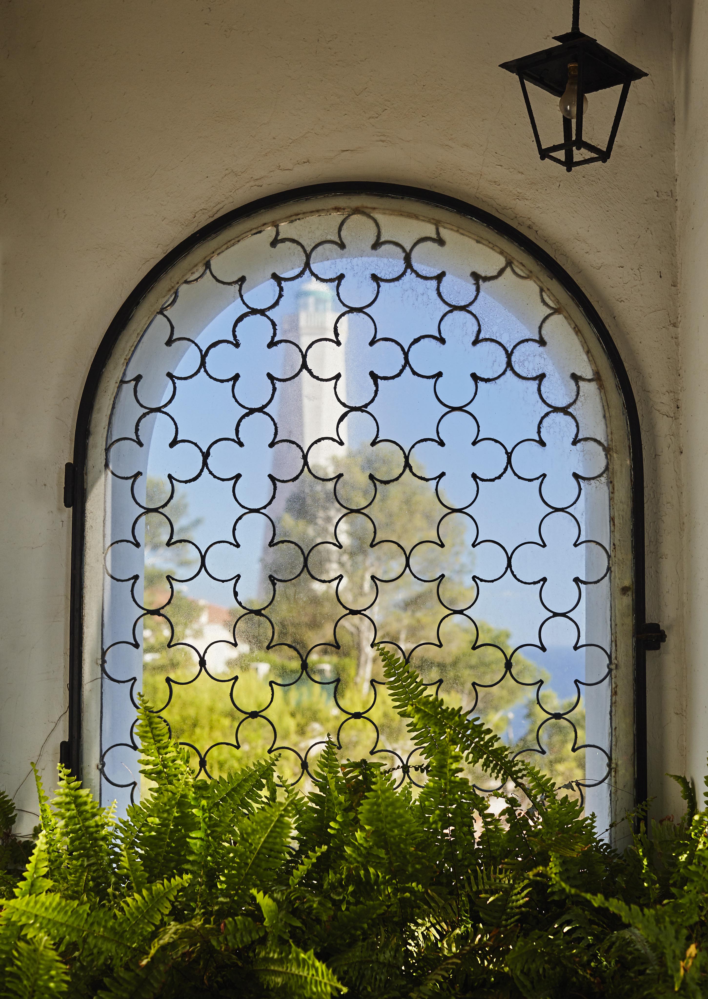 Arched window with decorative iron grille, overlooking trees and distant scenery. A lantern hangs above, and ferns are visible in the foreground.