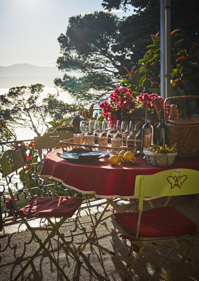 Table set for outdoor dining with glasses, wine, and fruit. Red tablecloth, surrounded by chairs, trees, and view of water in the background. Bright sunlight and plants nearby.