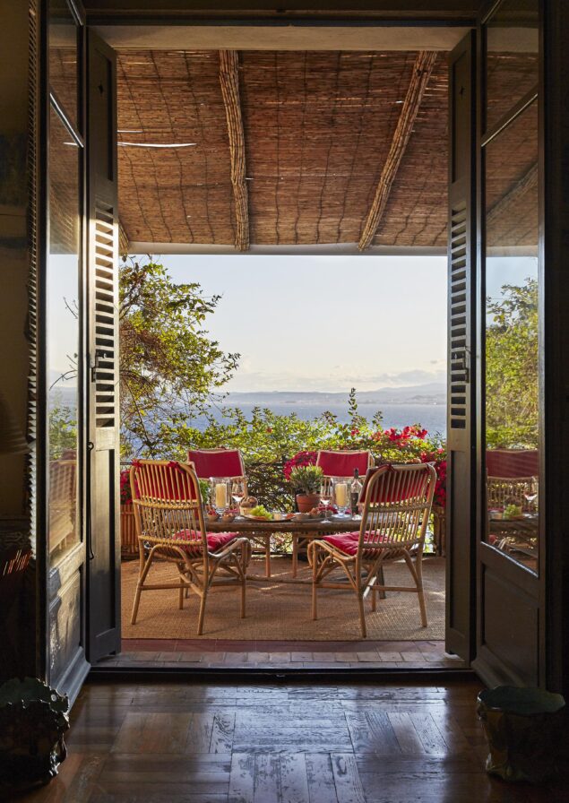 Open double doors lead to a cozy terrace with two wicker chairs and a table set for two, overlooking a scenic view surrounded by trees and flowers.