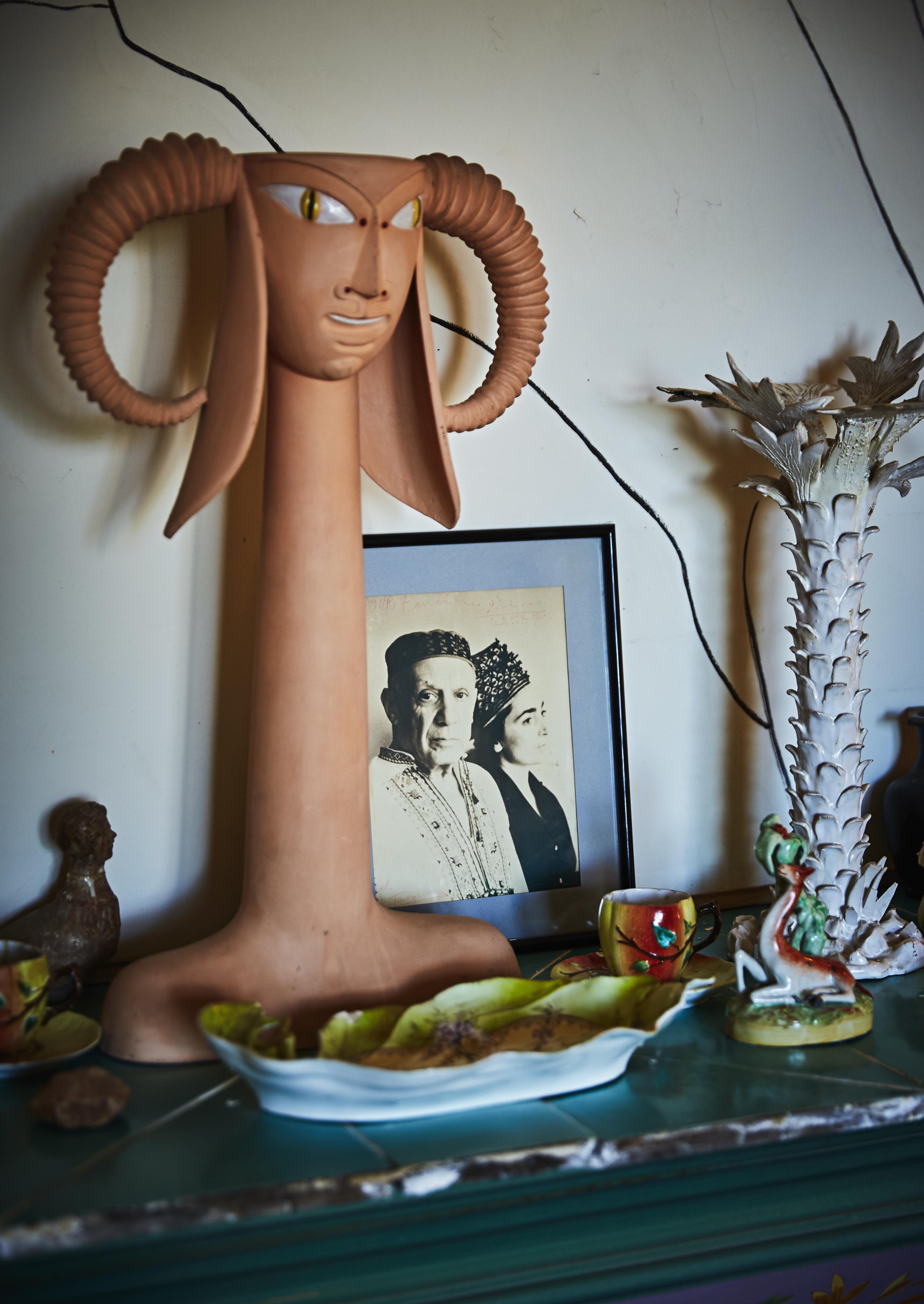 A ceramic sculpture with large horns is displayed on a shelf. Behind it, a framed vintage black-and-white photo is visible. Various decorative items and a dish with food are placed nearby.