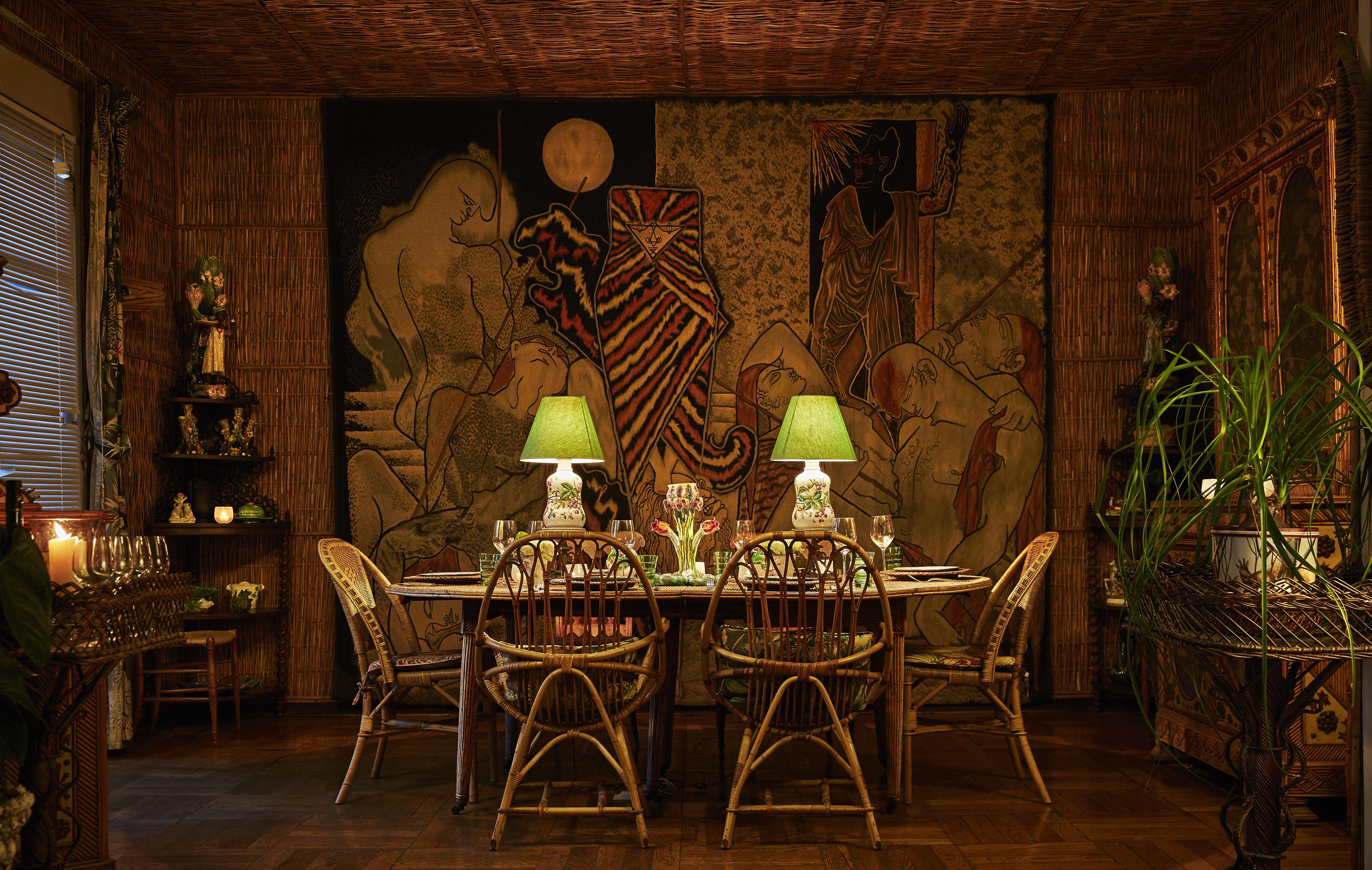 A dimly lit dining room with a wicker table and chairs, decorative table lamps with green shades, and a large, intricate tapestry on the wall.
