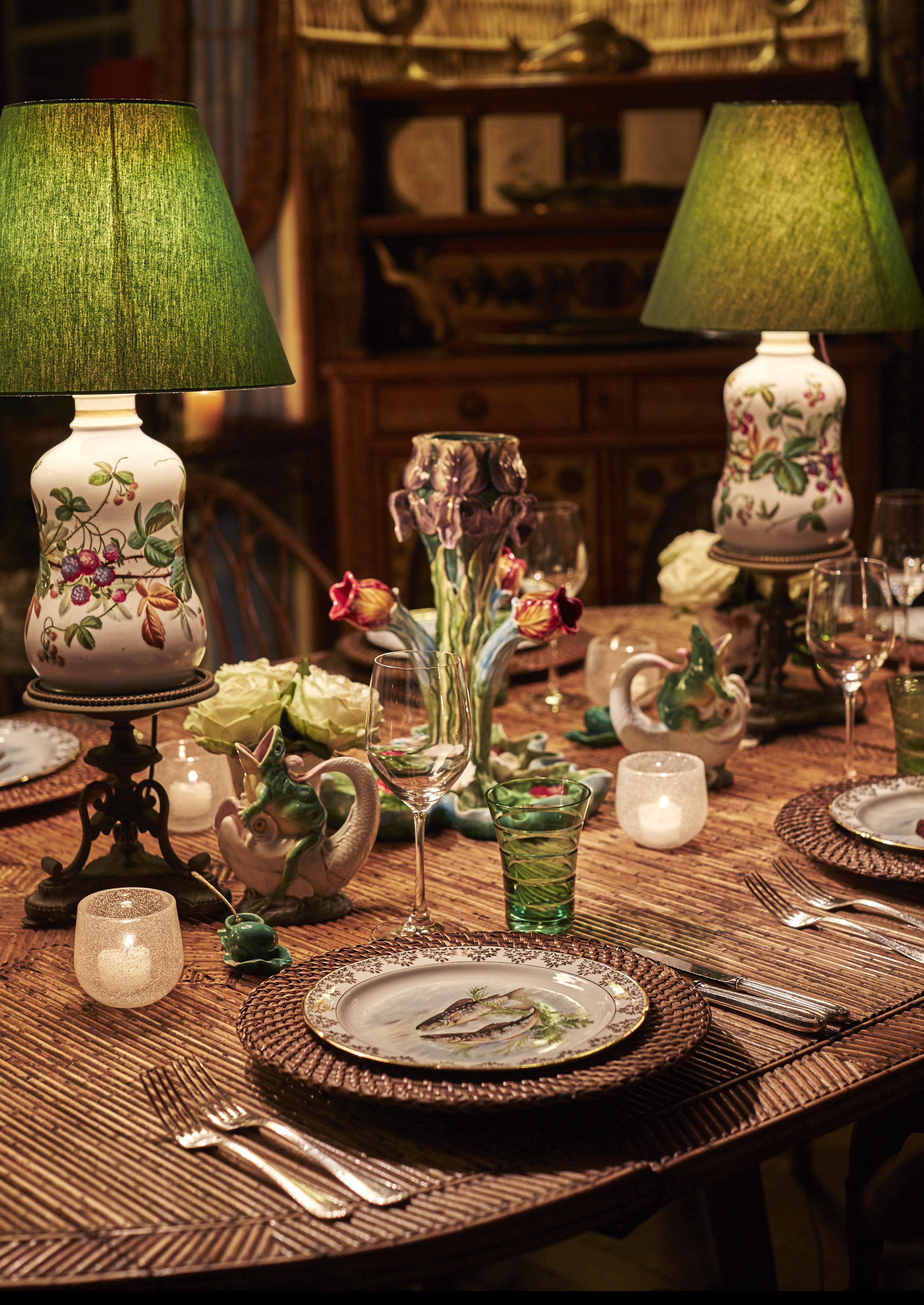 A dining table set with floral-patterned plates, glasses, candles, and an ornate vase. Two green-shaded lamps provide light.