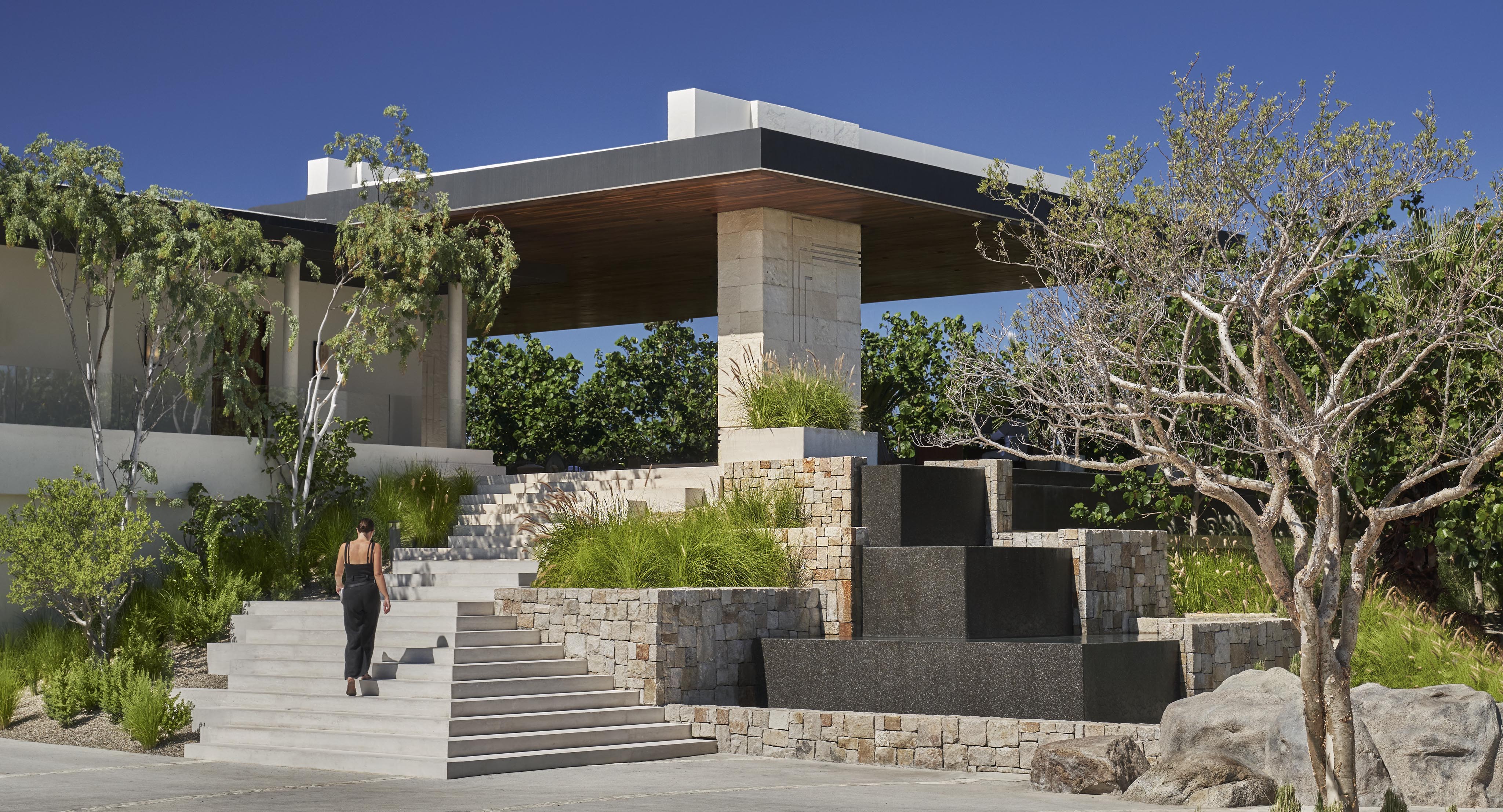 A person ascends the exterior steps of luxury Four Seasons Los Cabos architecture beside a modern building with stone and wood elements, lush greenery, and a cascade water feature, under a clear blue sky.