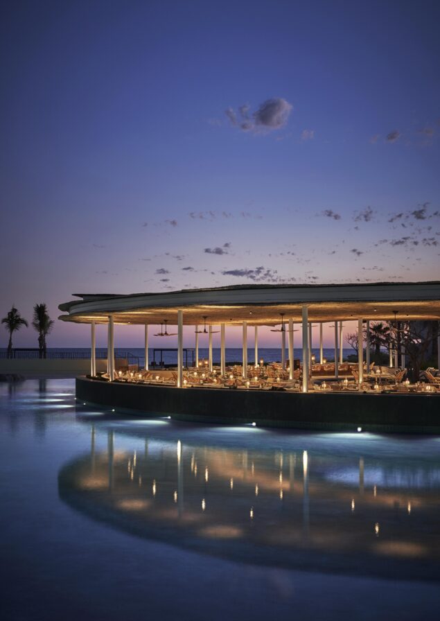 Exterior architecture of Four Seasons Los Cabos open air restaurant; Casa de Brasa, captured at evening, reflection in water and sky a deep blue graduating to a soft pink at the horizon.