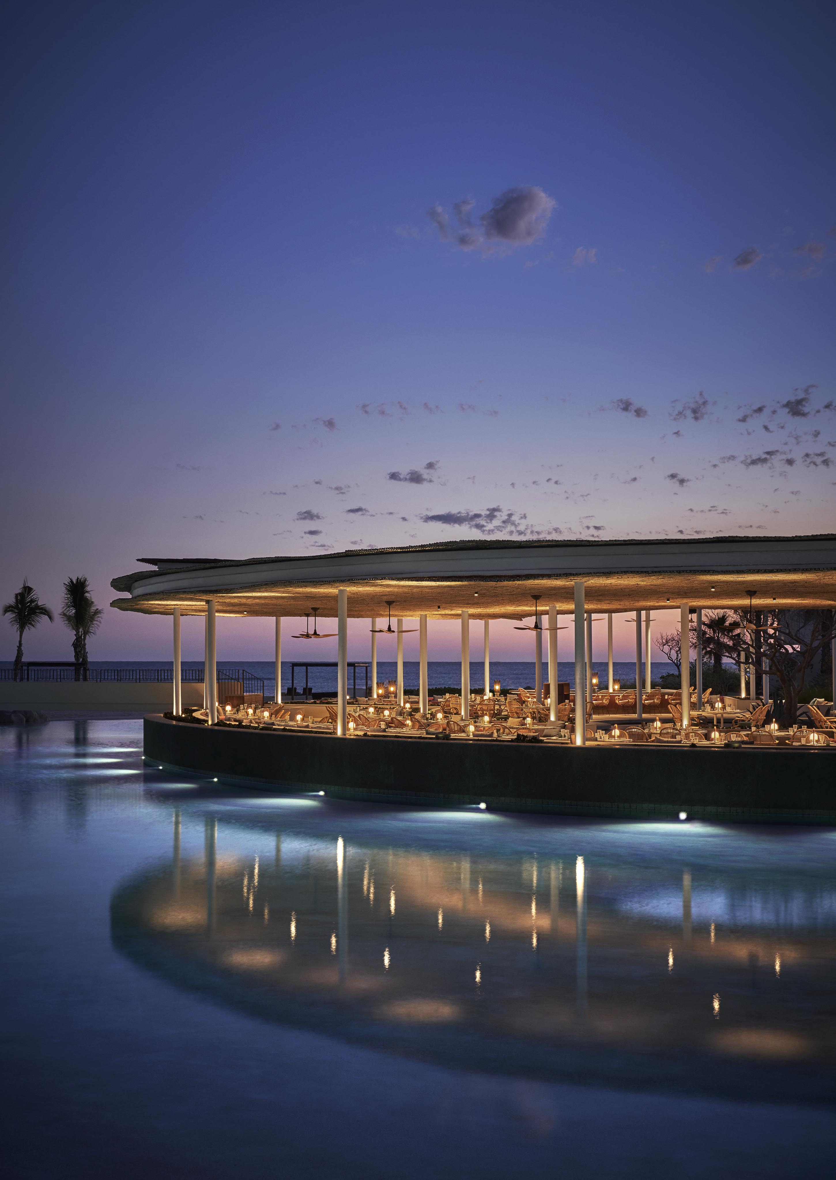 Exterior architecture of Four Seasons Los Cabos open air restaurant; Casa de Brasa, captured at evening, reflection in water and sky a deep blue graduating to a soft pink at the horizon.