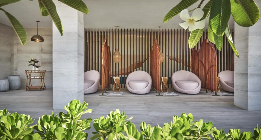 Three round chairs face surfboard-shaped wooden dividers in a modern outdoor Four Seasons Los Cabos at Costa Palmas private spa and lounge area, with lush green plants and white flowers in the foreground.