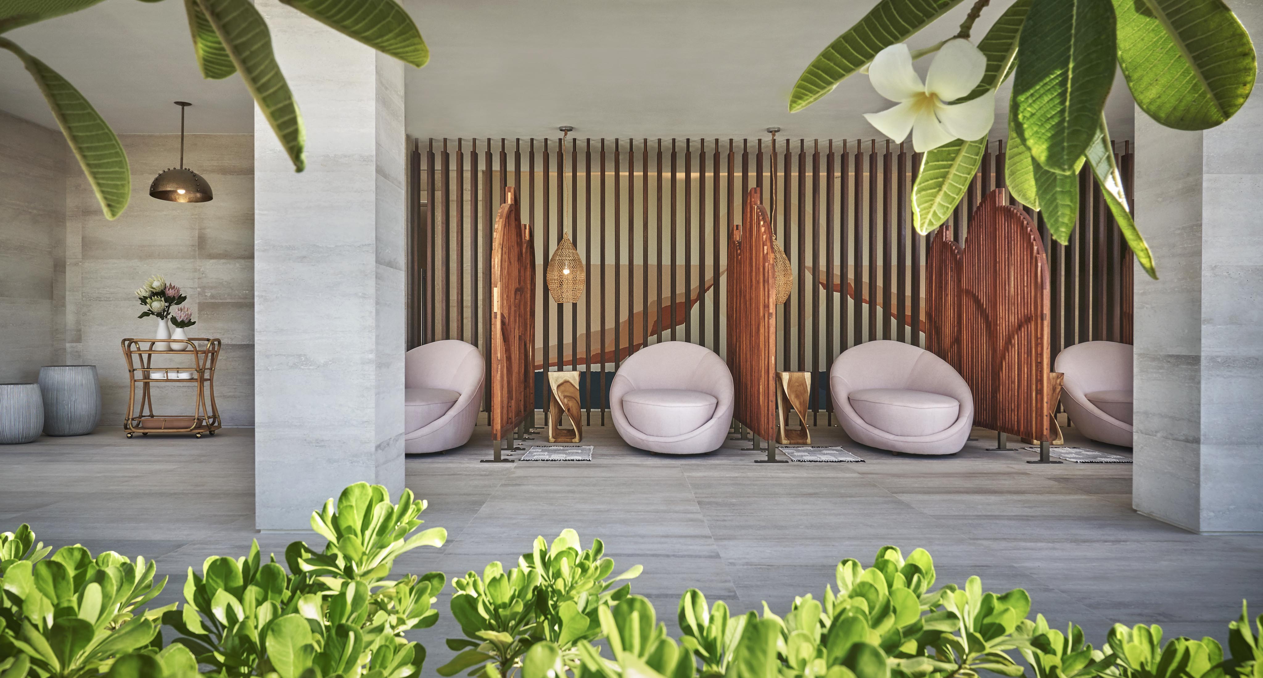 Three round chairs face surfboard-shaped wooden dividers in a modern outdoor Four Seasons Los Cabos at Costa Palmas private spa and lounge area, with lush green plants and white flowers in the foreground.