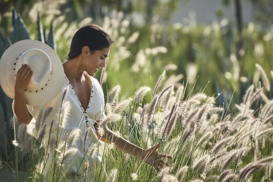 Woman wearing white flowing dress and hat walks through pampas grass and sunlight near the Four Seasons Los Cabos at Costa Palmas spa exterior