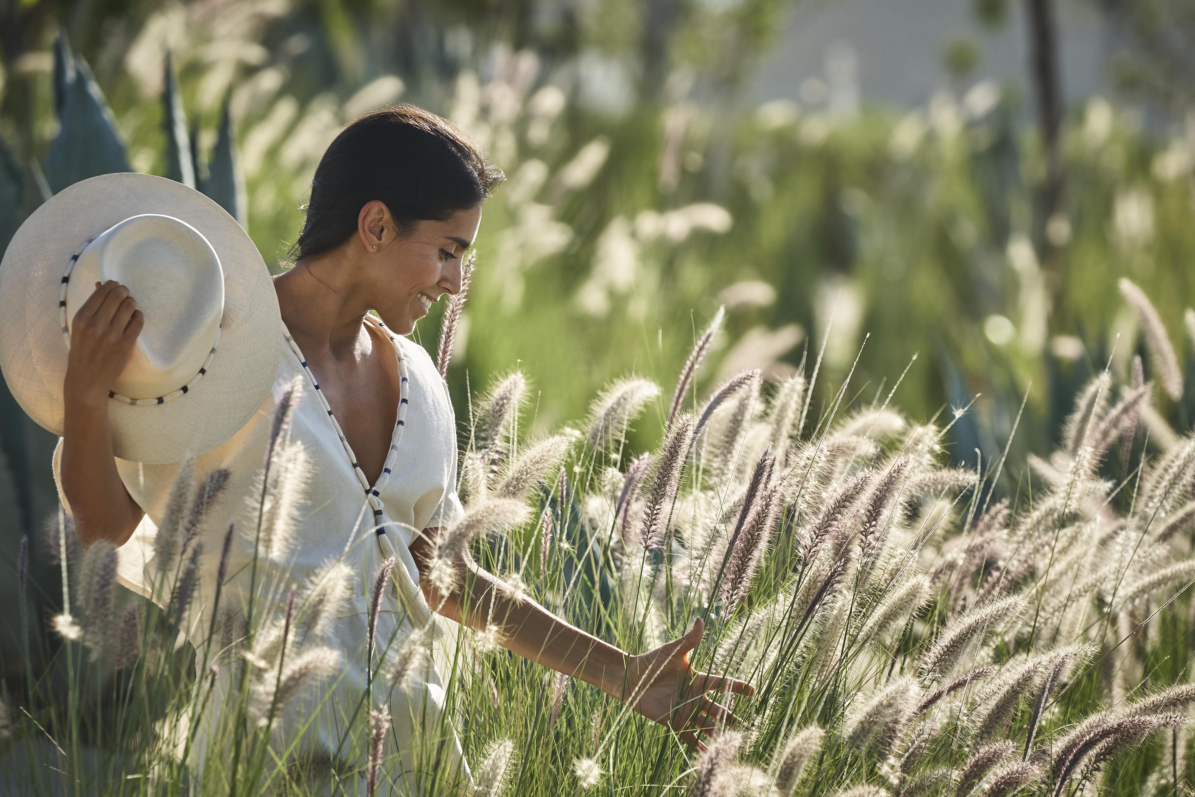 Woman wearing white flowing dress and hat walks through pampas grass and sunlight near the Four Seasons Los Cabos at Costa Palmas spa exterior