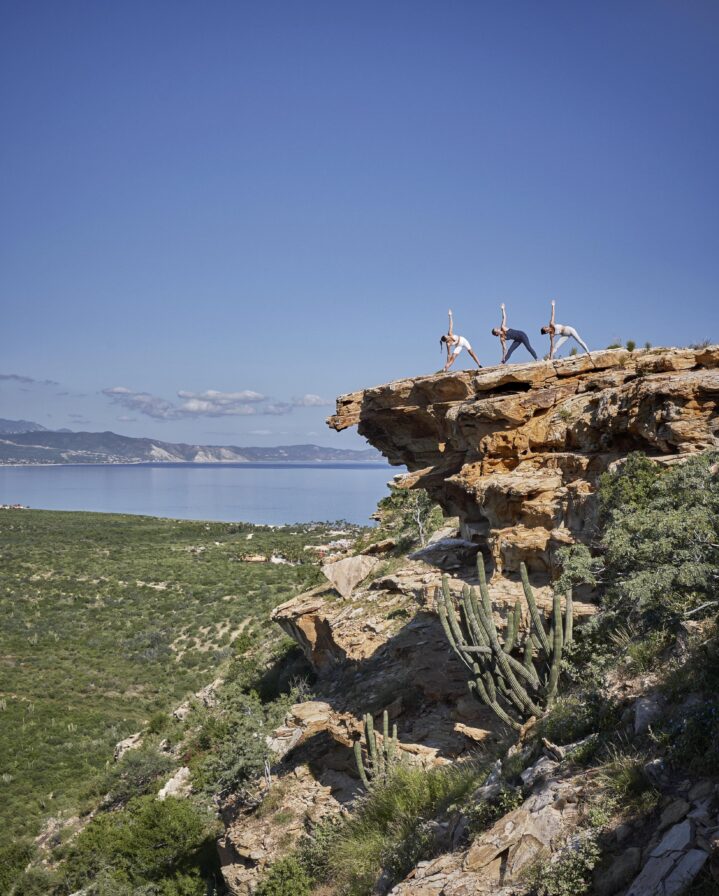Three people perform yoga poses on a rocky cliff of Los Cabos destination of flat rock; overlooking a vast landscape and ocean under a clear blue sky.