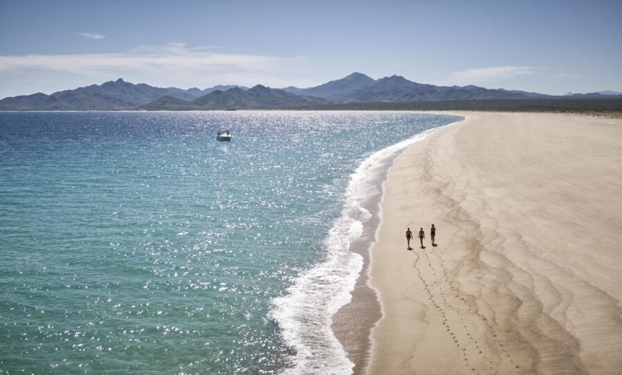 Three people walk along a private Costa Palmas beach near the shoreline, leaving footprints behind them, with mountains visible in the background.