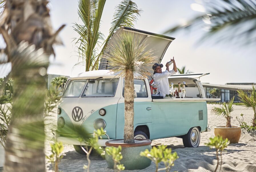 Joyous Four Seasons Los Cabos hospitality bar staff mixes cocktails within a converted van bar on a sandy beach, Palm trees are in the background.