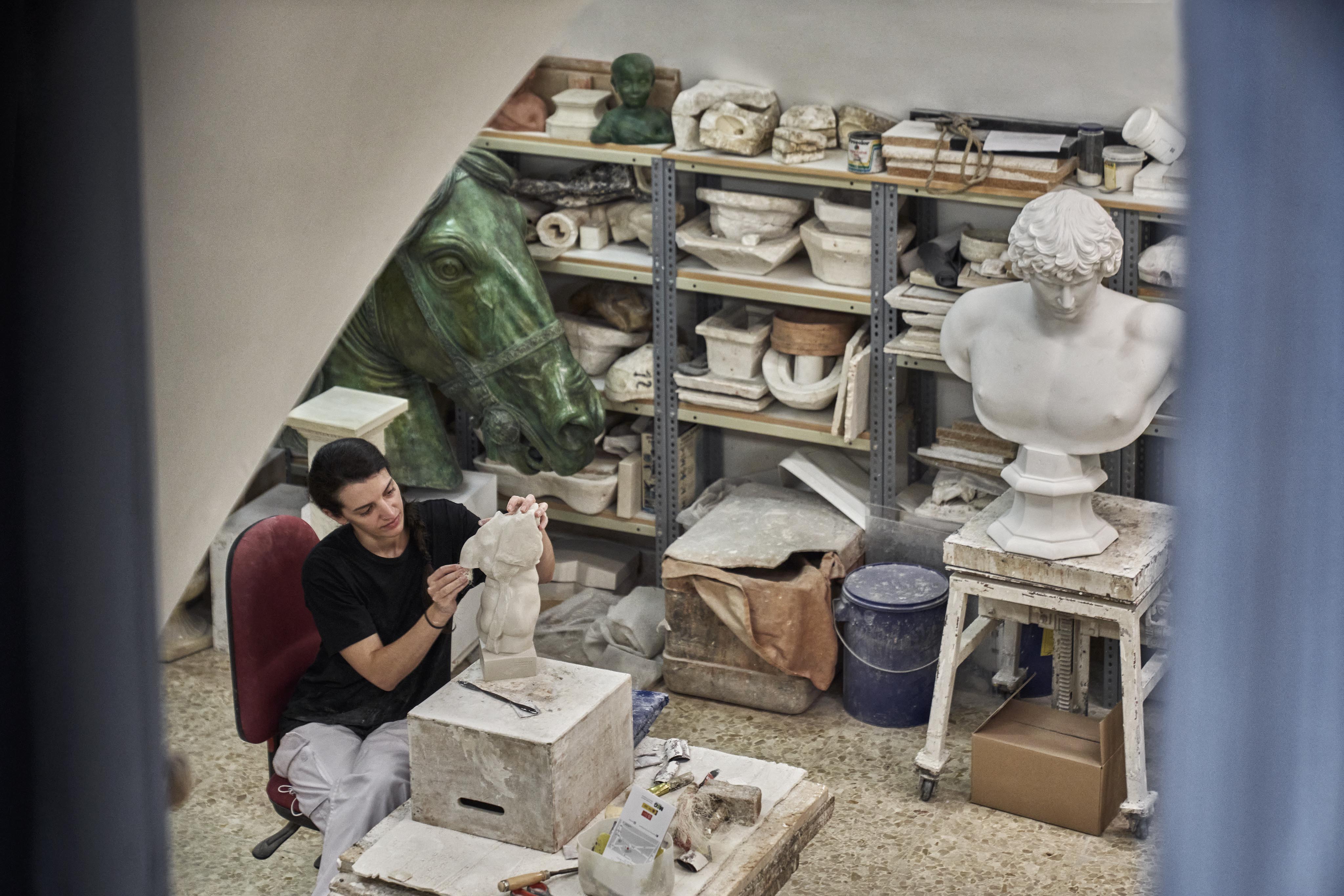 Person sculpting a small statue in a studio filled with various plaster casts and sculptures, including a bust and a horse head.