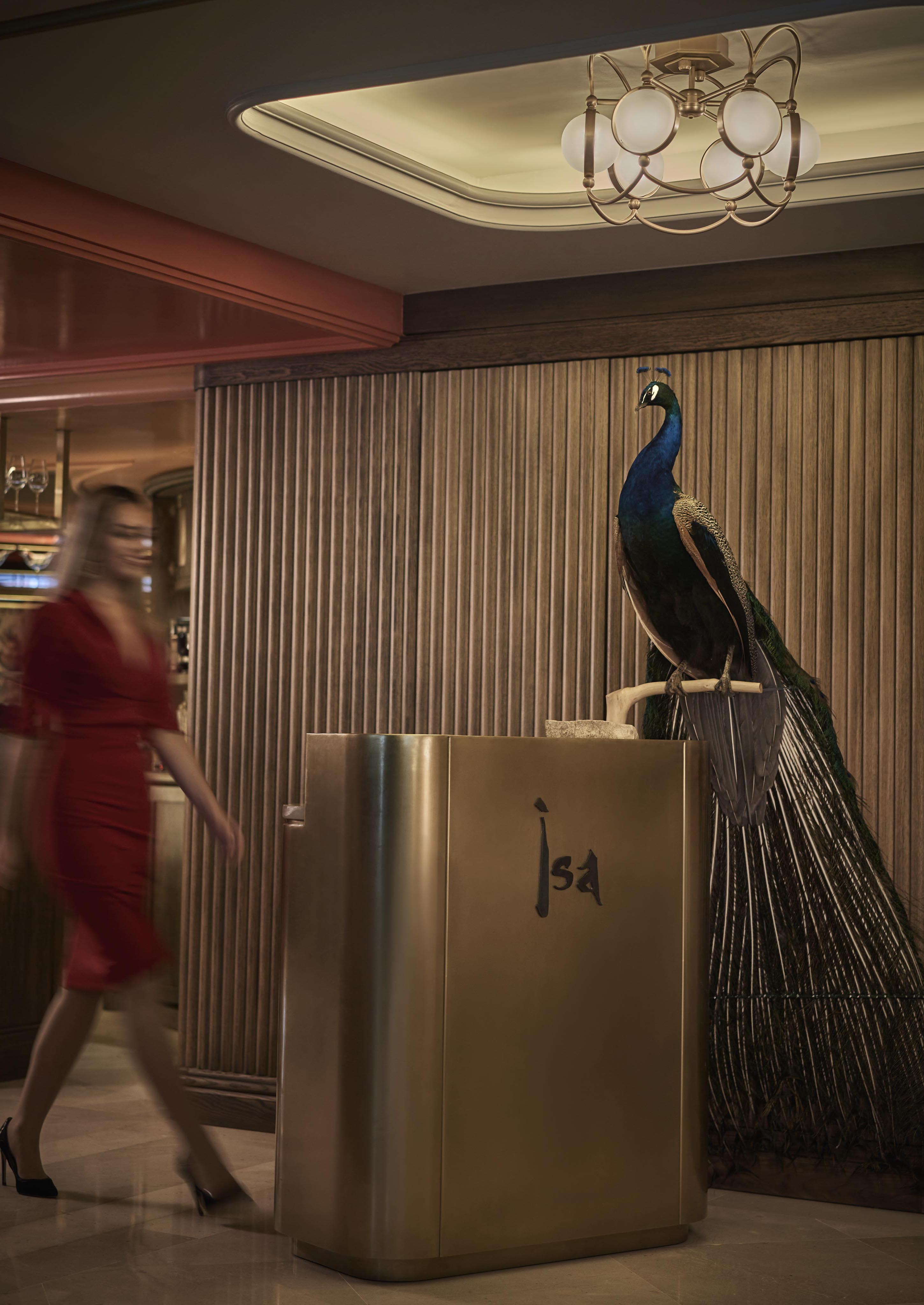 A woman in a red dress walks past a podium with the word "Issa" on it, featuring a large decorative peacock beside it.
