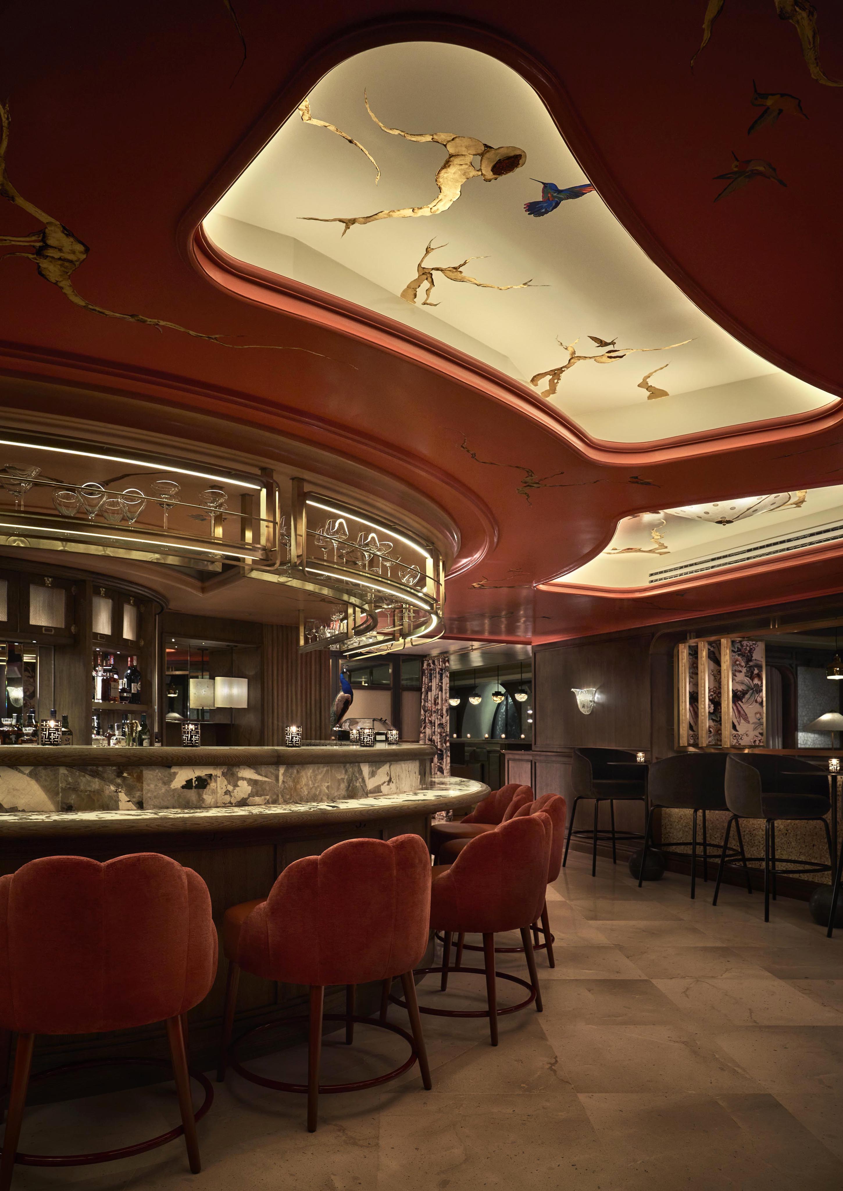 Elegant bar interior with a curved marble counter, plush red barstools, and a creatively designed ceiling featuring artistic patterns and soft lighting.