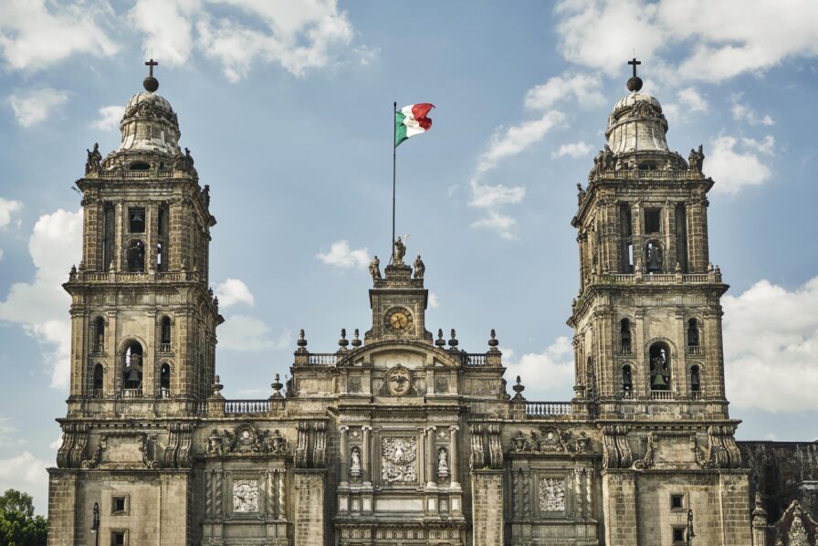 The Metropolitan Cathedral in Mexico City stands tall with a Mexican flag at its center, beneath a partly cloudy sky—an iconic scene captured by a destination travel photographer.