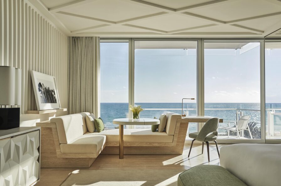 Modern Four Seasons Miami hotel room with beige and green decor features a seating area, round table, large windows, and balcony overlooking the ocean. Sunlight illuminates the space.