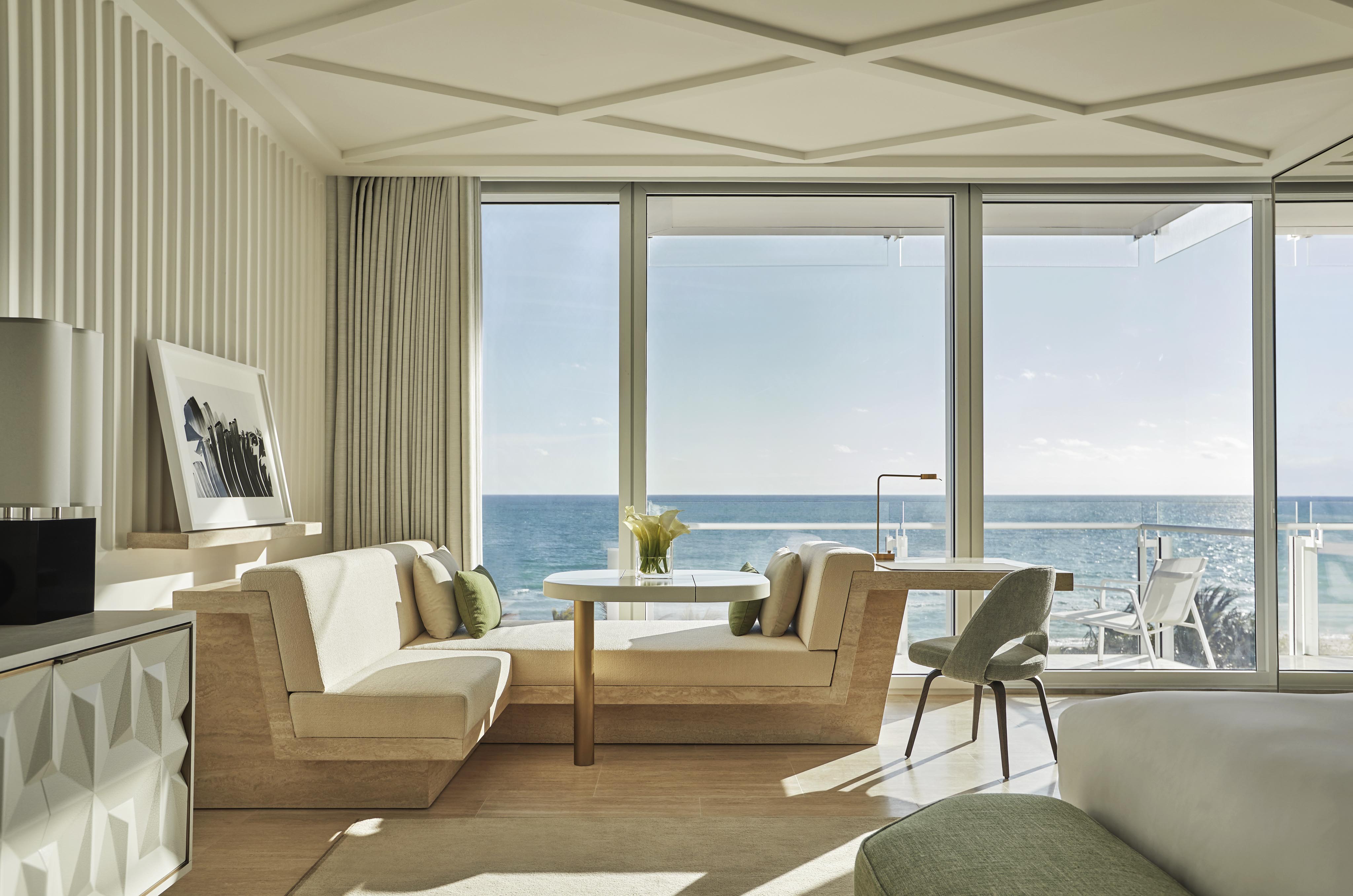 Modern Four Seasons Miami hotel room with beige and green decor features a seating area, round table, large windows, and balcony overlooking the ocean. Sunlight illuminates the space.