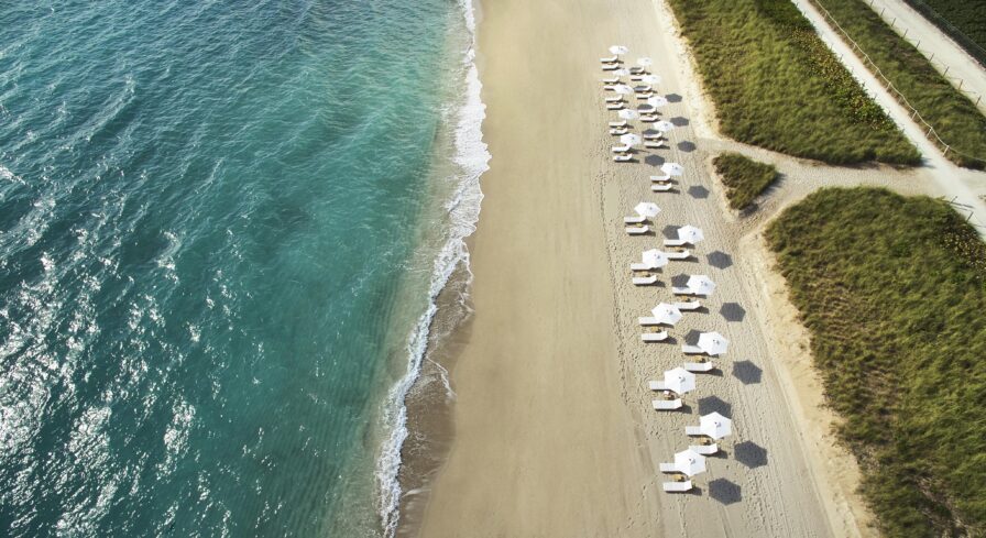 Aerial view of a pristine Miami Florida beach with rows of white Four Seasons Surf Club resort umbrellas and sun loungers along the sandy shore. Green vegetation borders the area on the right.