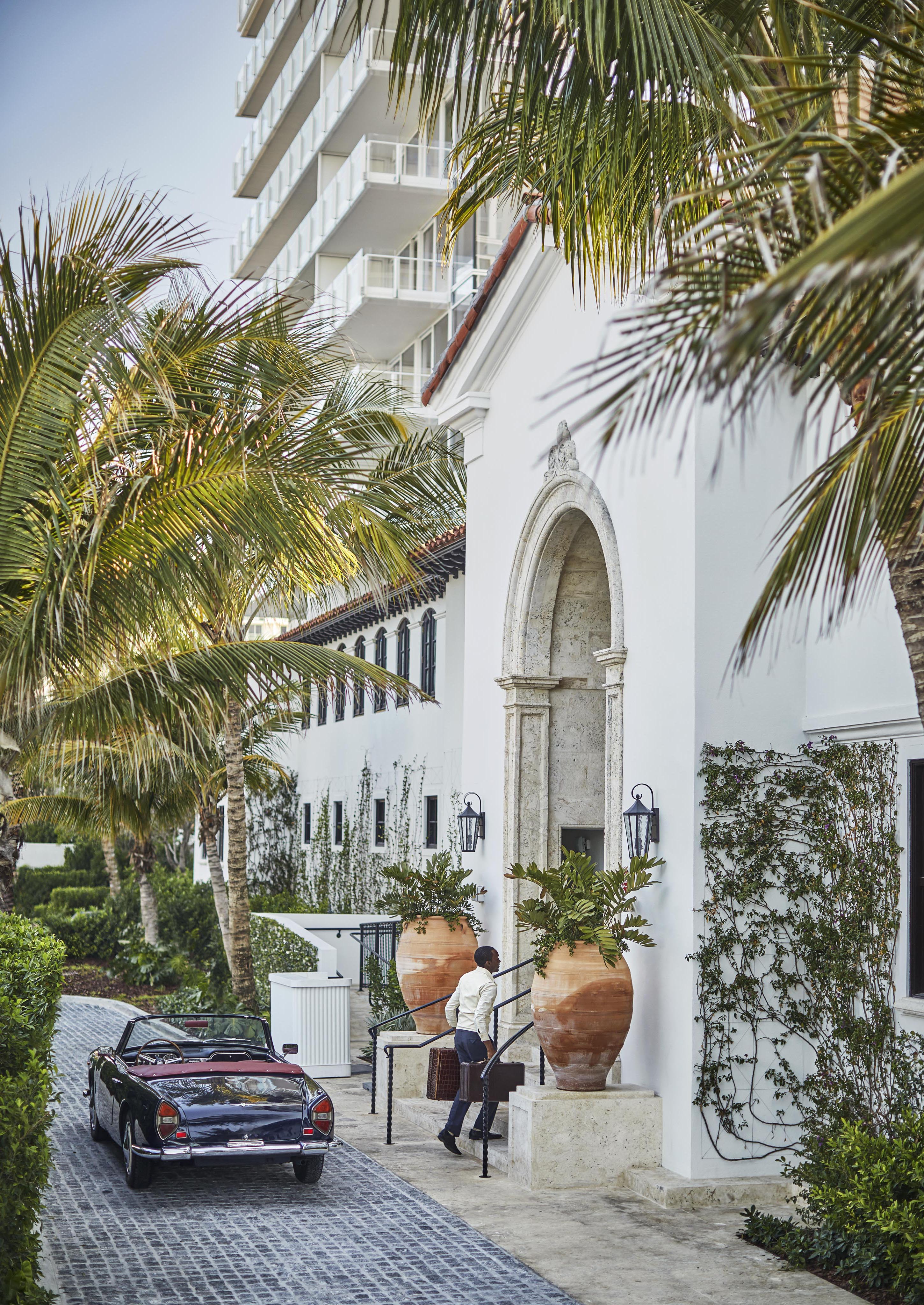 Hospitality staff walks into Four Seasons Miami Surf Club lobby interior entrance, featuring a white building with large planters. A convertible car is parked on the cobblestone driveway, surrounded by palm trees.