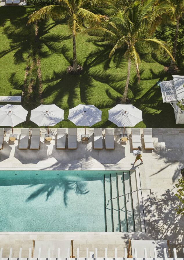 Aerial view of a exclusive Four Seasons Surf Club Miami outdoor resort pool area with luxury lounge chairs and umbrellas. A hospitality staff member walks beside the pool carrying towels, surrounded by shaded grass and greenery.
