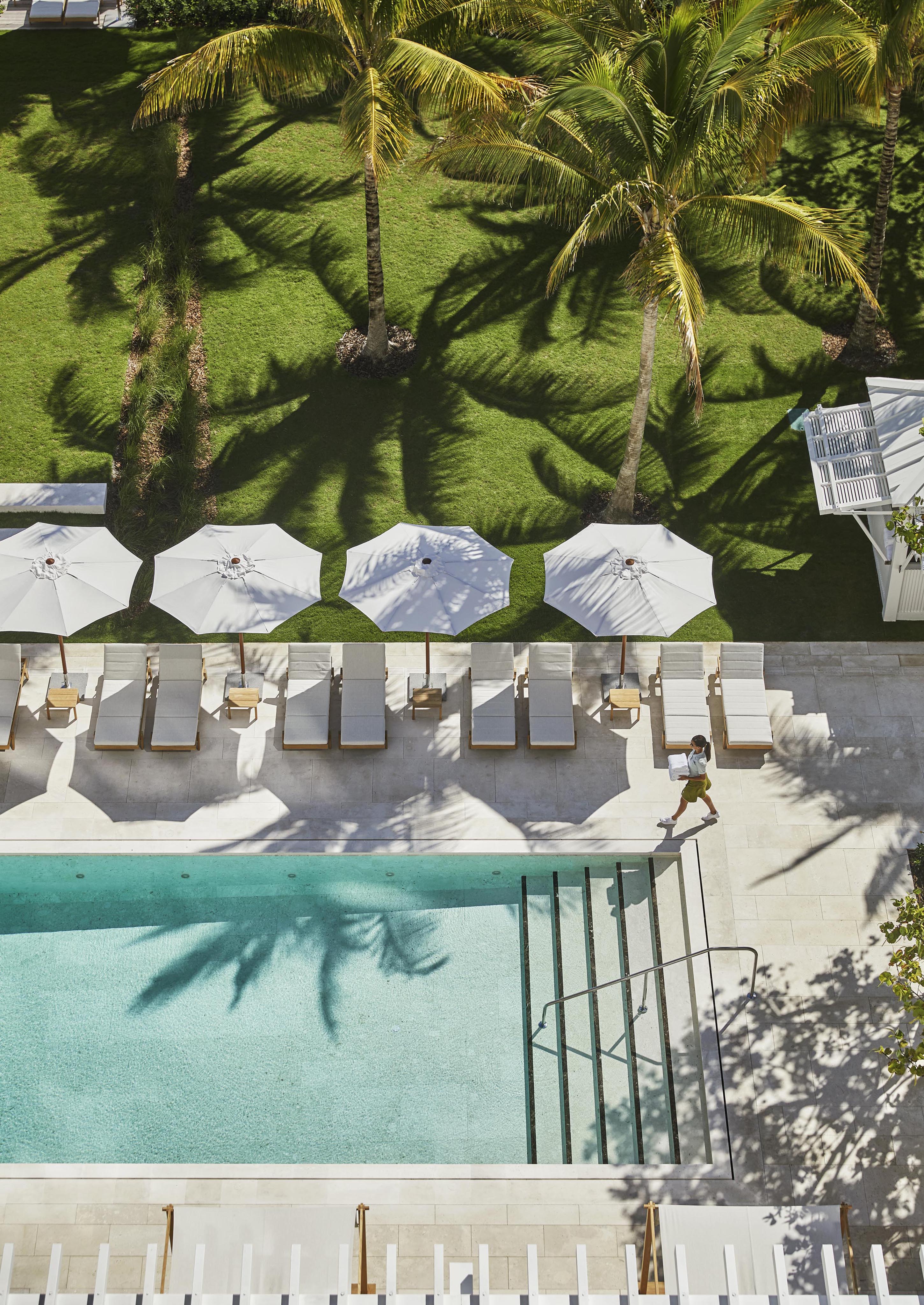 Aerial view of a exclusive Four Seasons Surf Club Miami outdoor resort pool area with luxury lounge chairs and umbrellas. A hospitality staff member walks beside the pool carrying towels, surrounded by shaded grass and greenery.