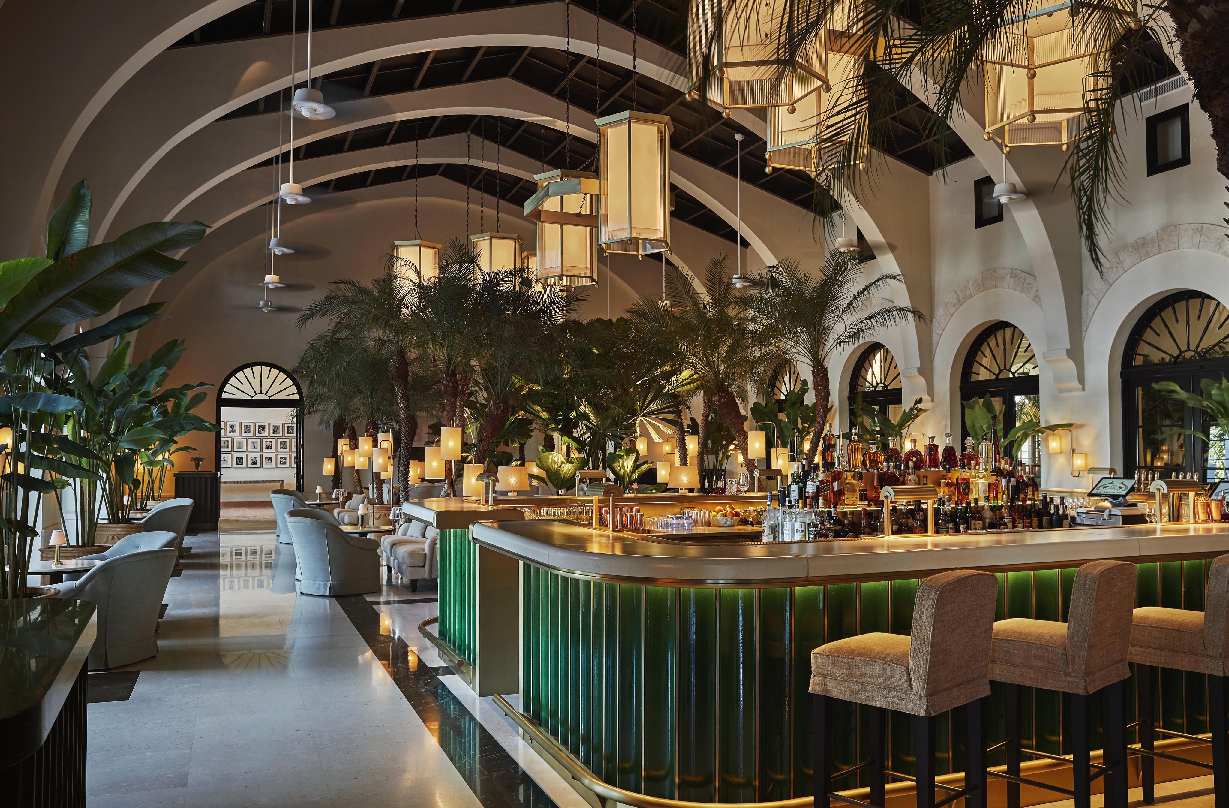 A Four Seasons Surf Club Champagne bar with a green-lit counter, high stools, tall lush Miami plants, arched windows, and modern hanging lights, set in an elegant interior with high ceilings.