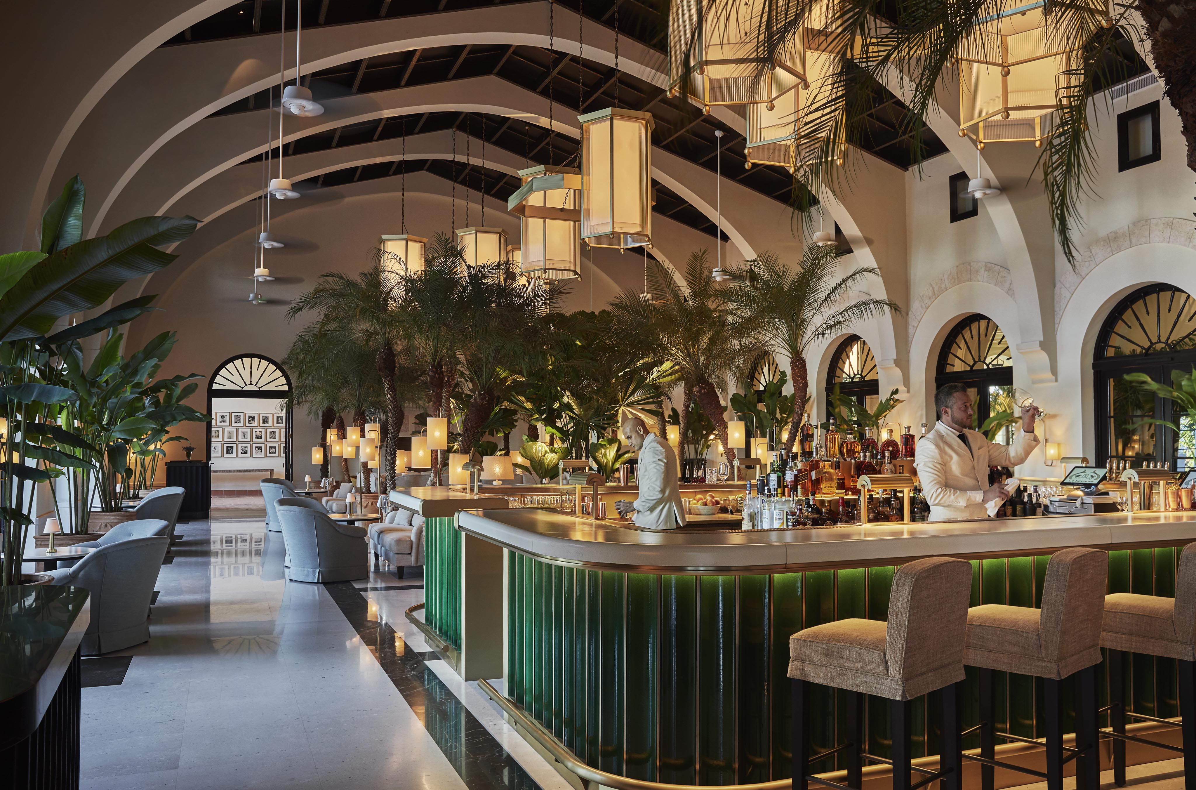 Elegant Four Seasons Surf Club Miami Champagne bar interior with arched ceilings, large hanging lights, and palm décor. Bartenders behind a green counter. Plush seating and a modern, airy atmosphere.