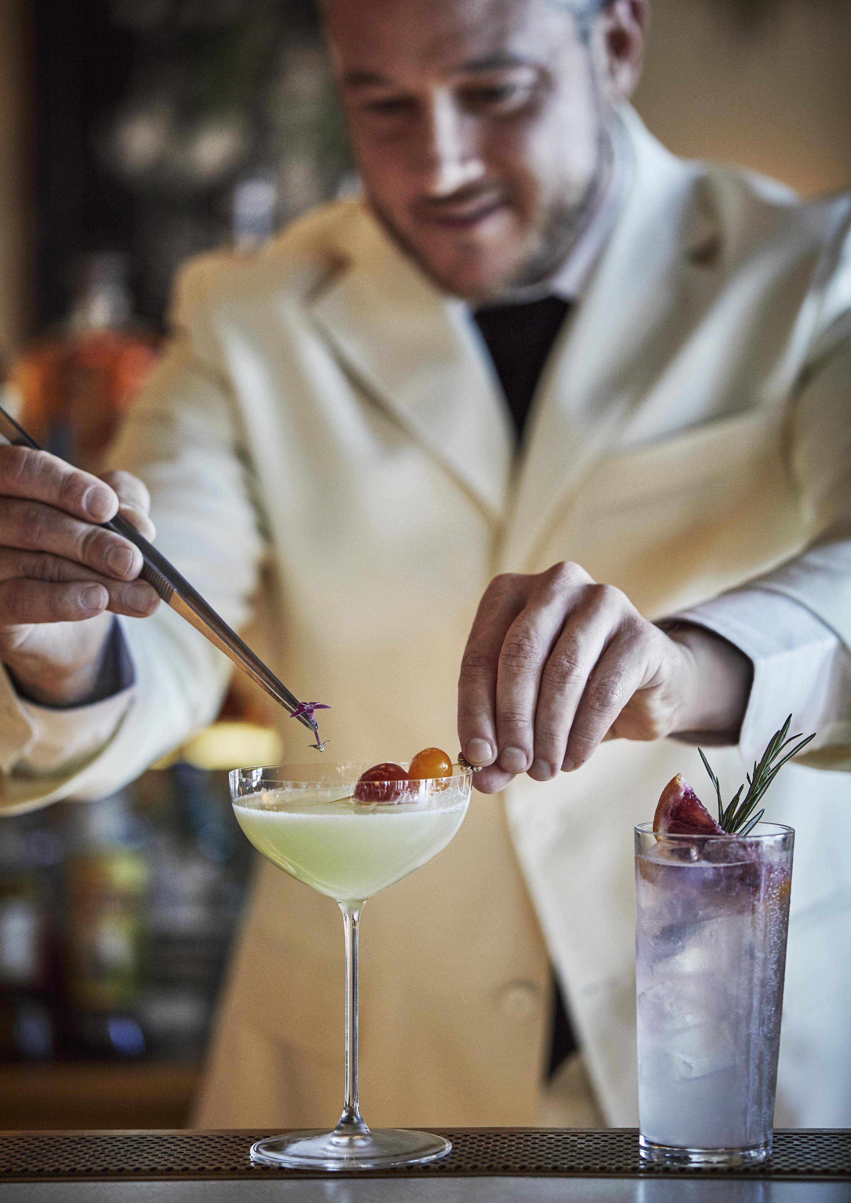 A Four Seasons Surf Club Miami resort bartender in a white jacket garnishes a yellow cocktail with a red cherry and adds a sprig to another drink in a tall glass with ice and purple fruit.