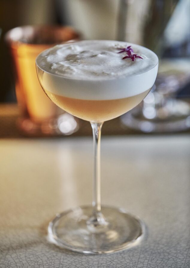 A cocktail glass filled with a frothy beverage, garnished with a small purple flower, set on a light-colored surface.