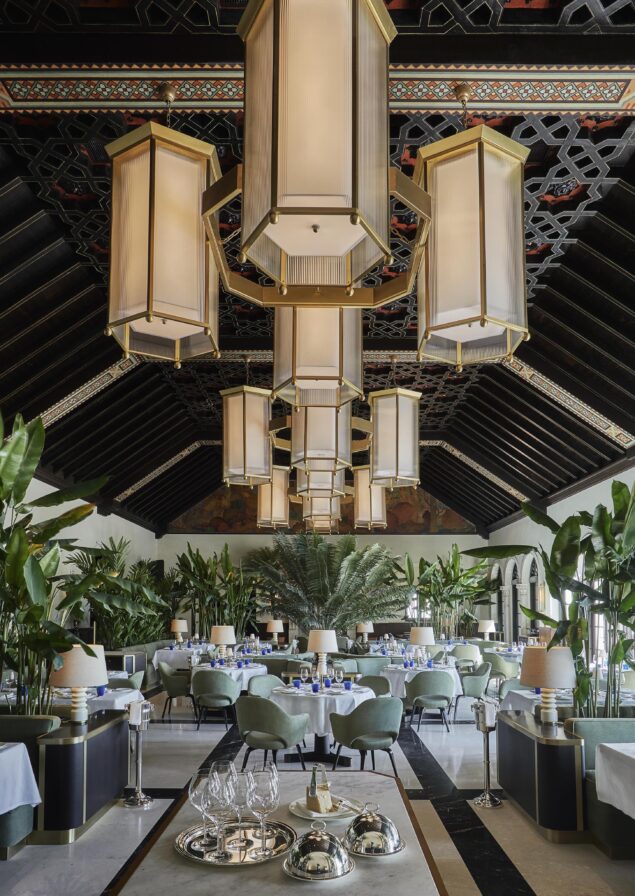 Opulent restaurant interior of Four Seasons Surf Club Miami with high wooden ceiling, large hanging lights, green plants, and neatly arranged tables with blue glasses and white linens.