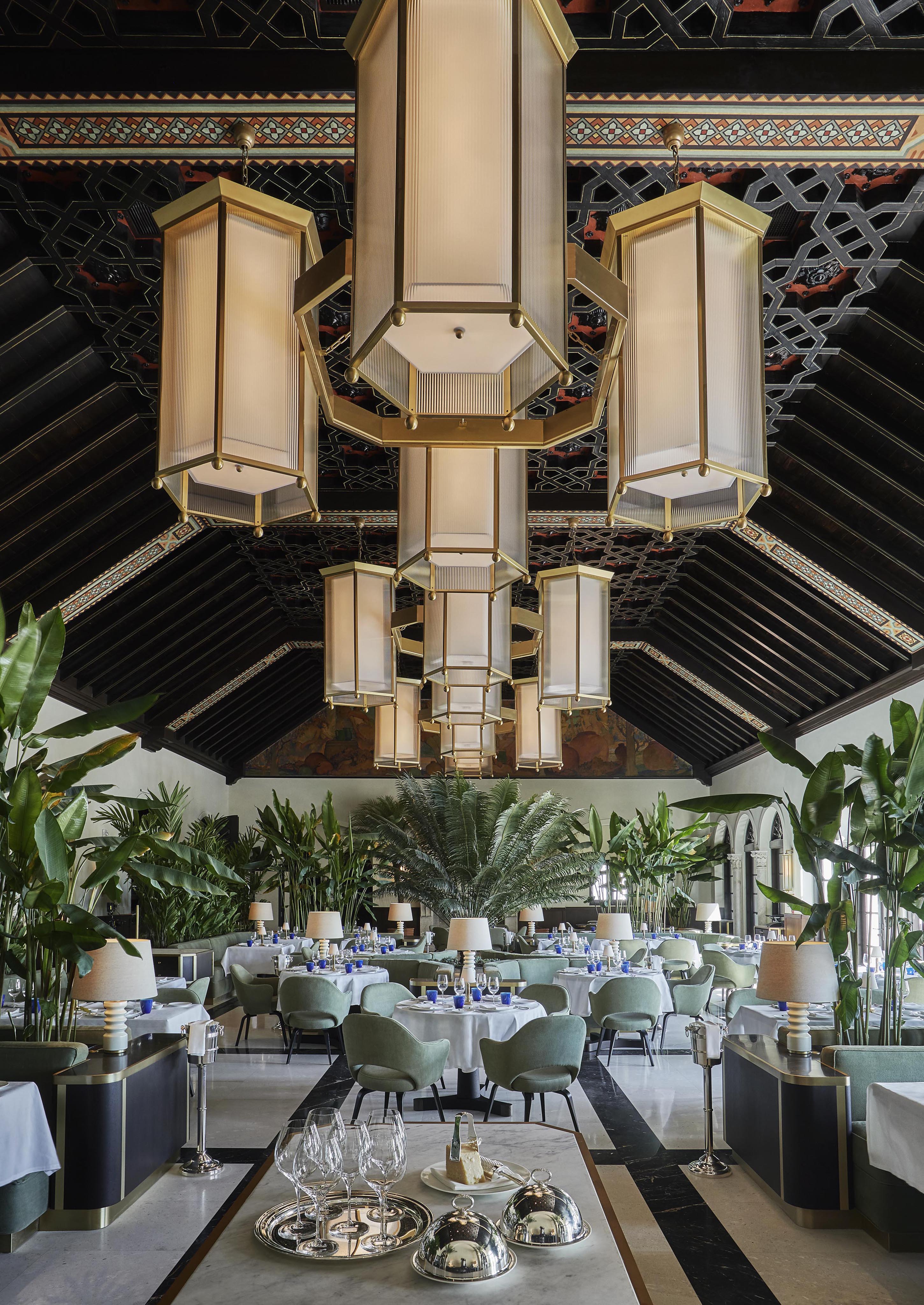 Opulent restaurant interior of Four Seasons Surf Club Miami with high wooden ceiling, large hanging lights, green plants, and neatly arranged tables with blue glasses and white linens.