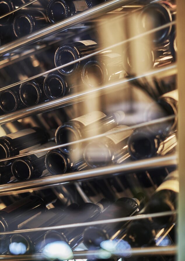 Rows of wine bottles stored horizontally on metal racks, seen through glass, in a wine cellar or store.