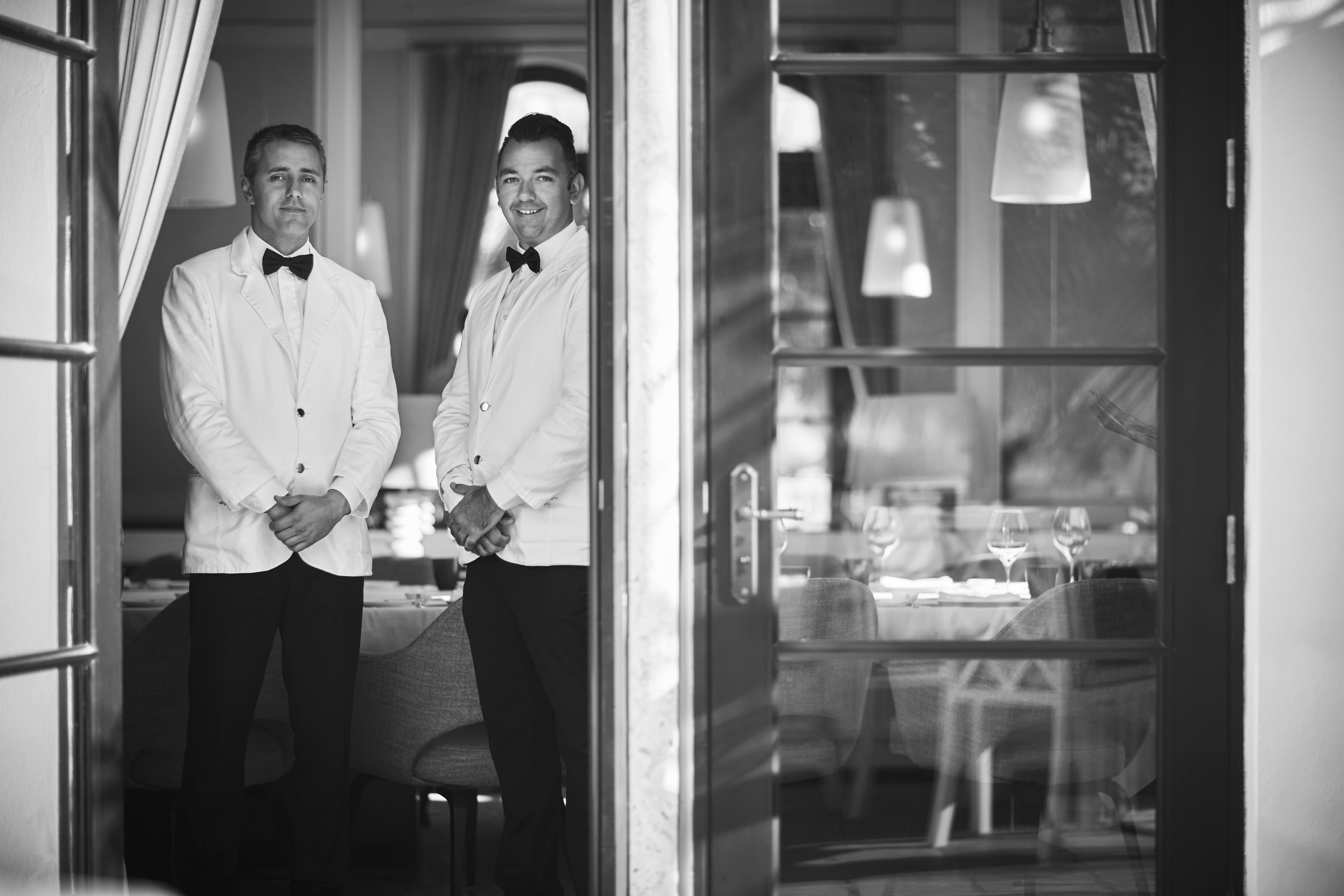 Two servers in white jackets and bow ties stand inside a restaurant by a glass door, with tables and chairs visible in the background.