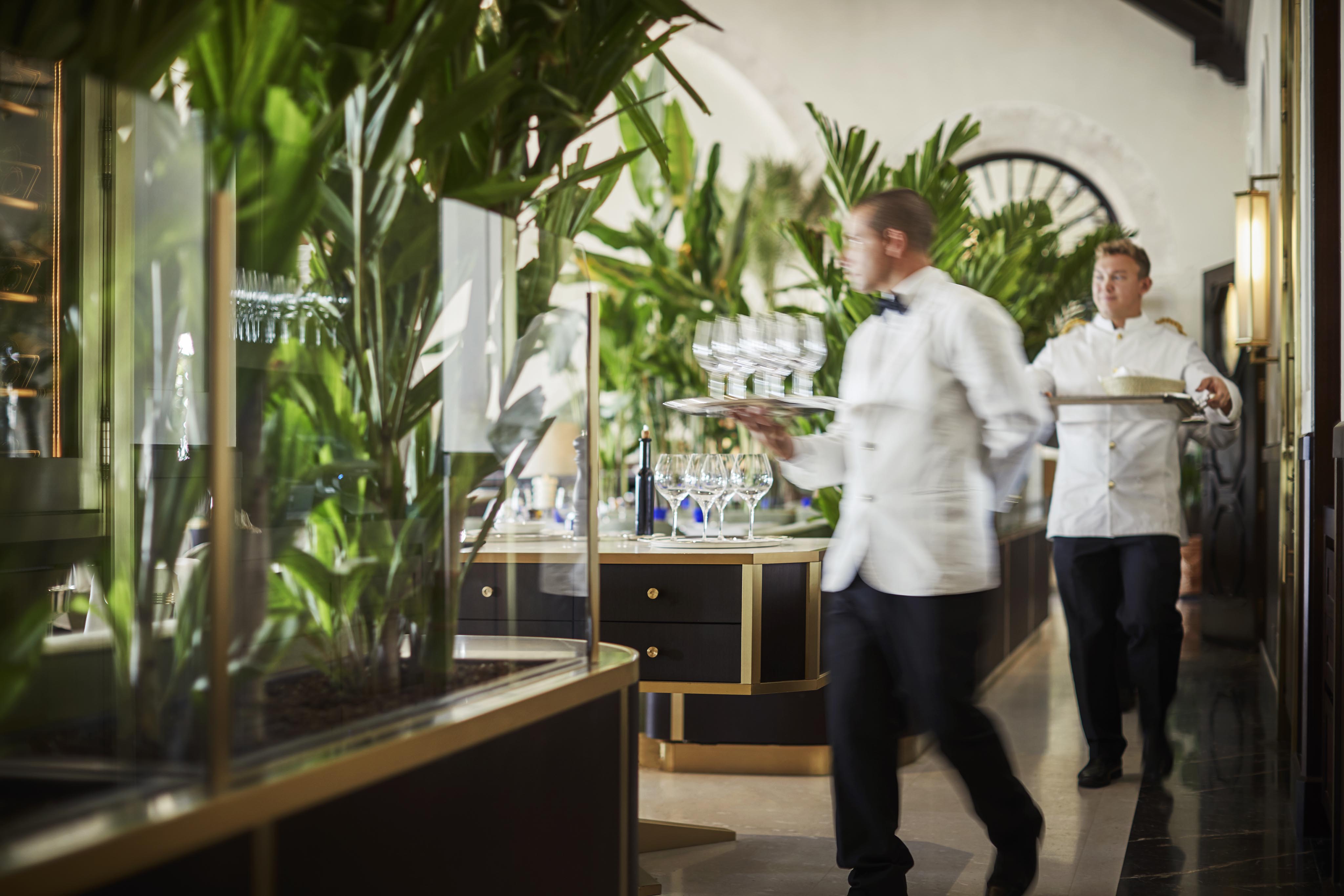Two Four Seasons Miami Surf Club hospitality staff in white uniforms carry trays of glassware through a restaurant with lush greenery and elegant interior.