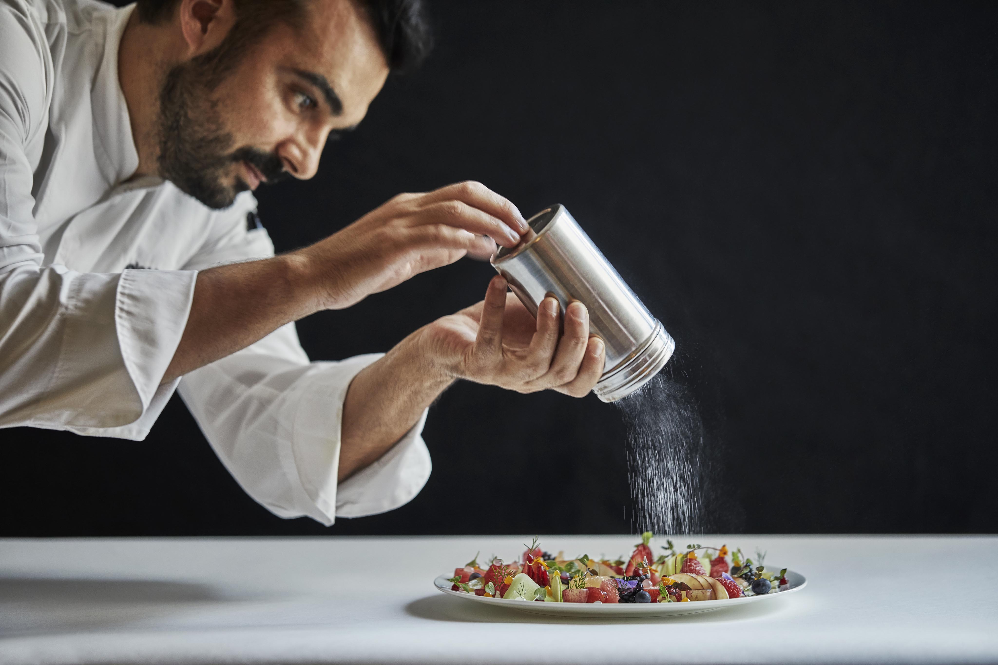 A Four Seasons Surf Club Miami resort chef sprinkles a fine powder over a plated dish on a white tablecloth, set against a dark background.