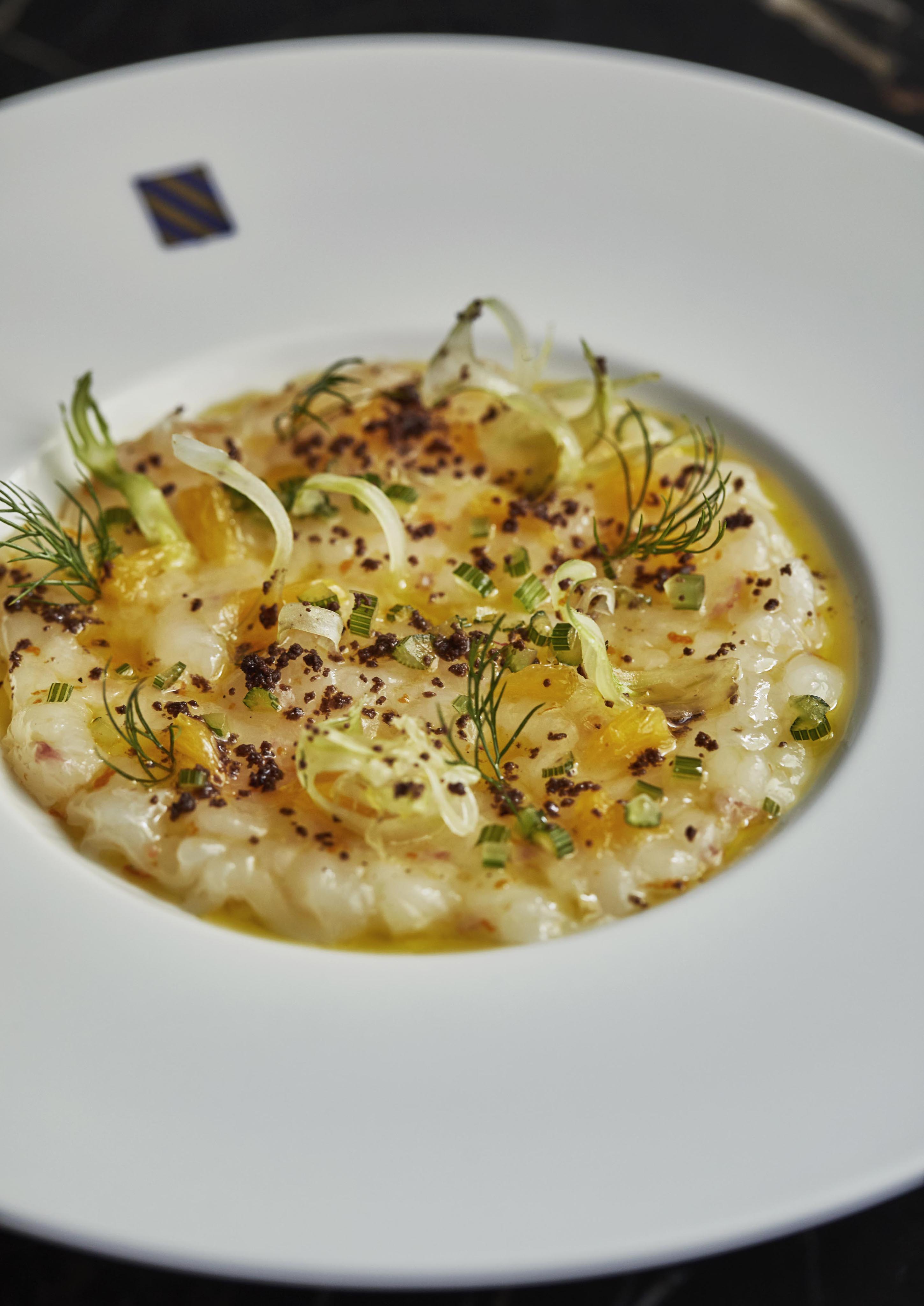 A plated dish of creamy risotto garnished with herbs, thinly sliced vegetables, and sprinkled spices, presented on a white plate with a subtle logo.