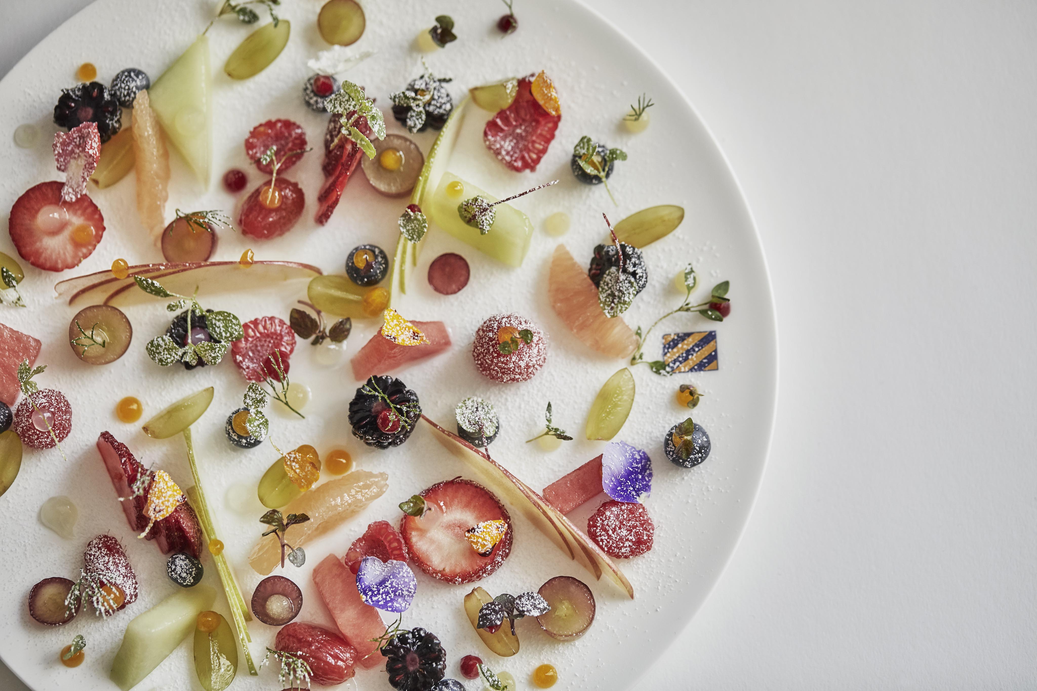 A white plate with an artistic arrangement of assorted fruits, edible flowers, and garnishes. Fruits include strawberries, melons, and berries, all lightly dusted with powdered sugar.