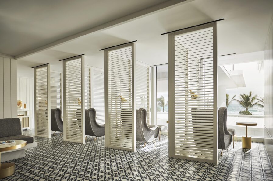 A minimalist Four Seasons Surf Club Miami luxury resort lounge area with patterned tile flooring, gray high-back chairs, and white slatted dividers. Large windows reveal palm trees and ocean views outside.