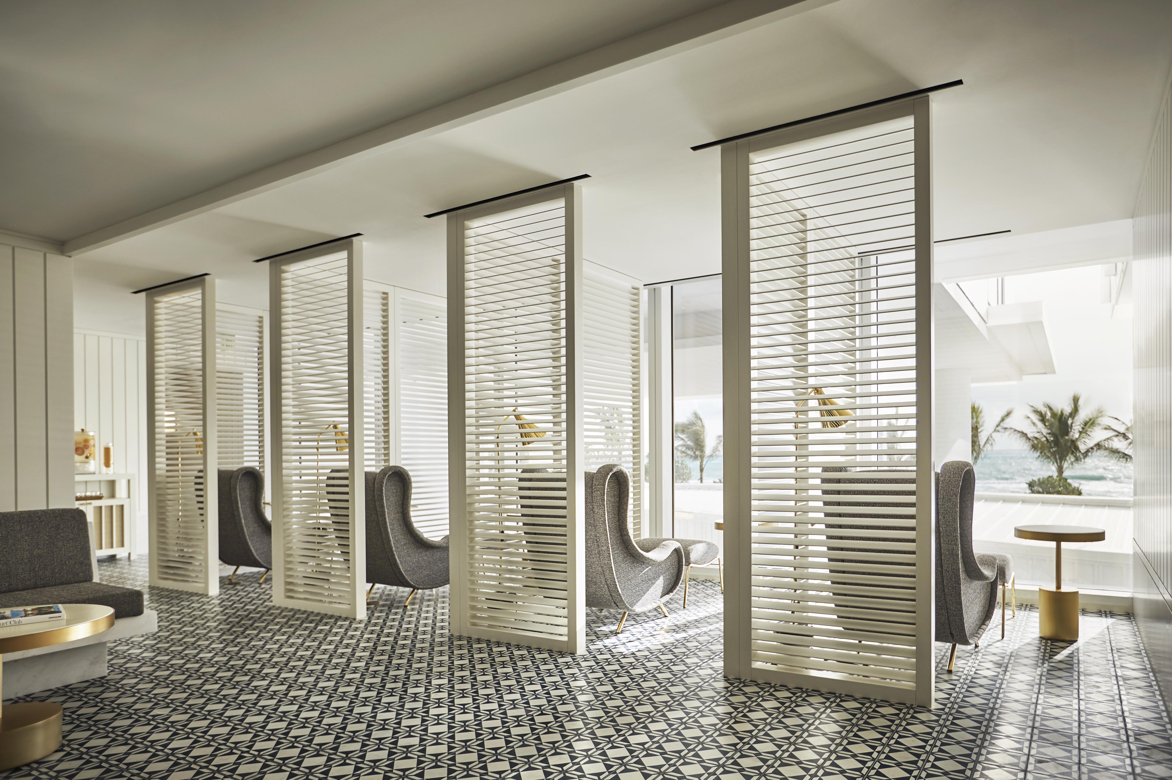 A minimalist Four Seasons Surf Club Miami luxury resort lounge area with patterned tile flooring, gray high-back chairs, and white slatted dividers. Large windows reveal palm trees and ocean views outside.