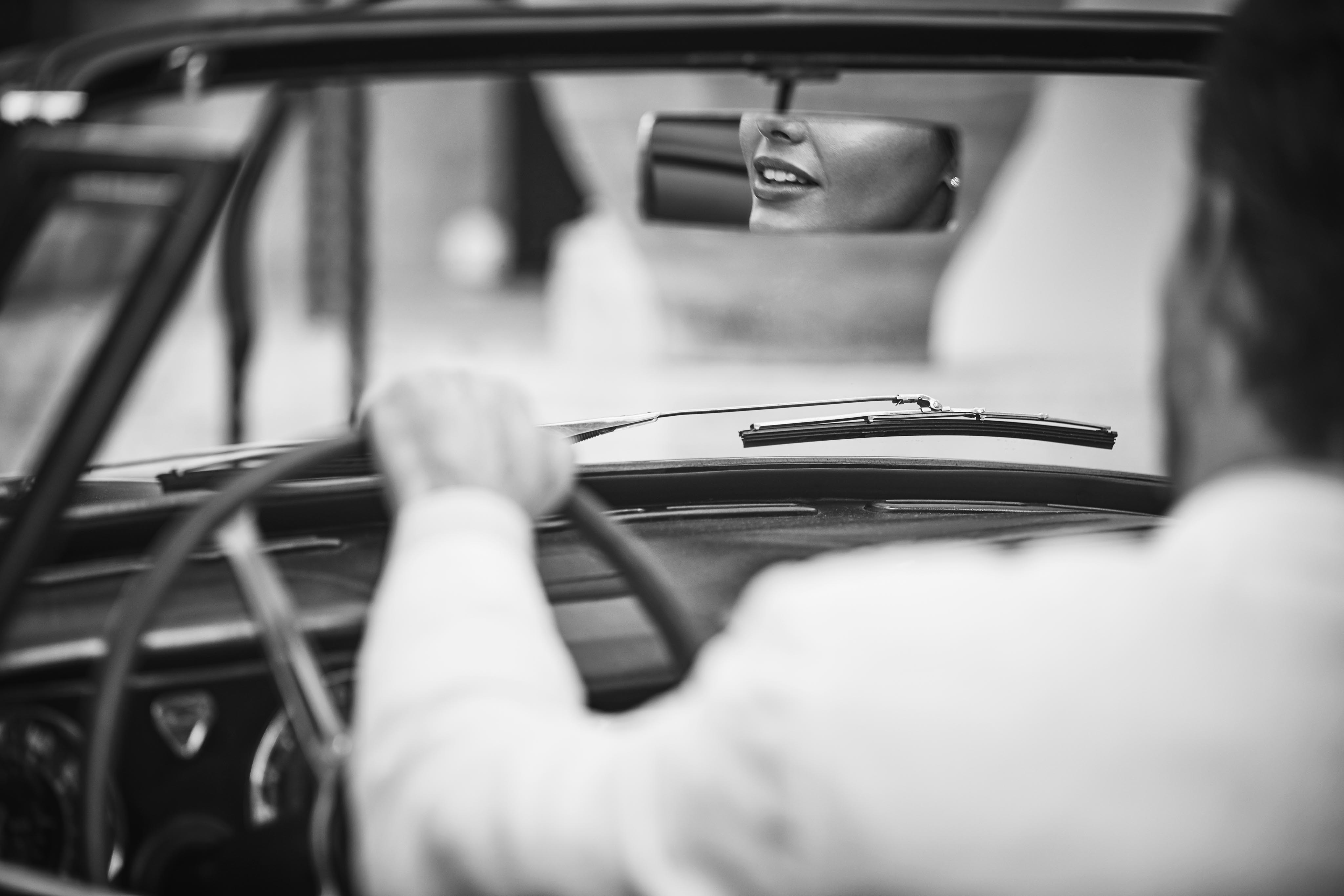 Black and white image of a person driving a convertible, viewed from behind. A passenger's reflection is visible in the rear-view mirror.