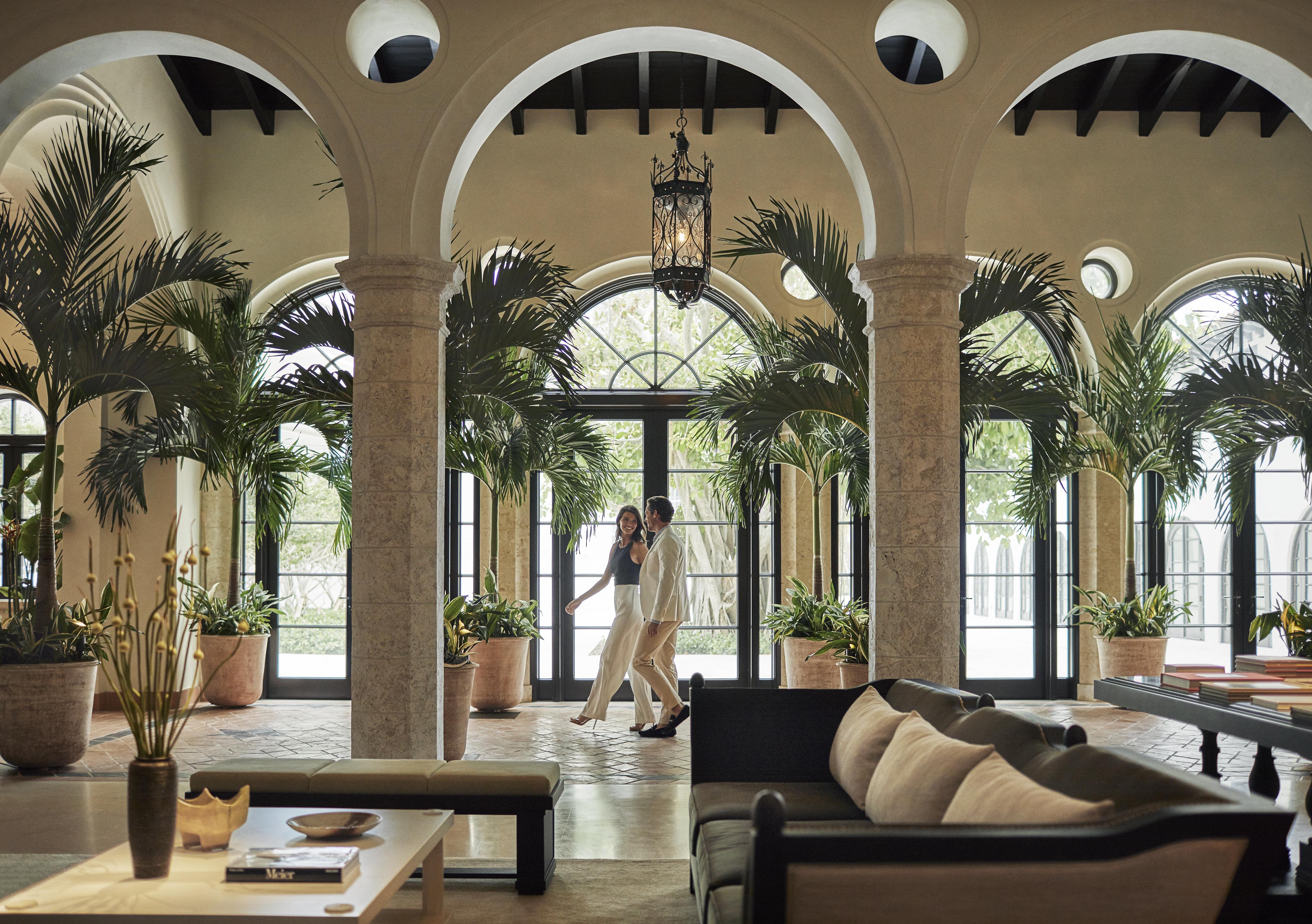 A couple walks hand in hand through a spacious, elegant Four Seasons Surf Club Miami hotel lobby with large windows, tall potted plants, and arched columns.
