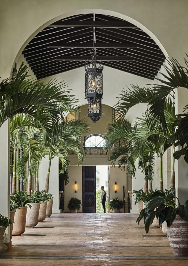A Four Seasons Surf Club Miami corridor with potted palm trees and a hanging lantern. Hospitality staff stands at the end near a large door, with sunlight streaming in.
