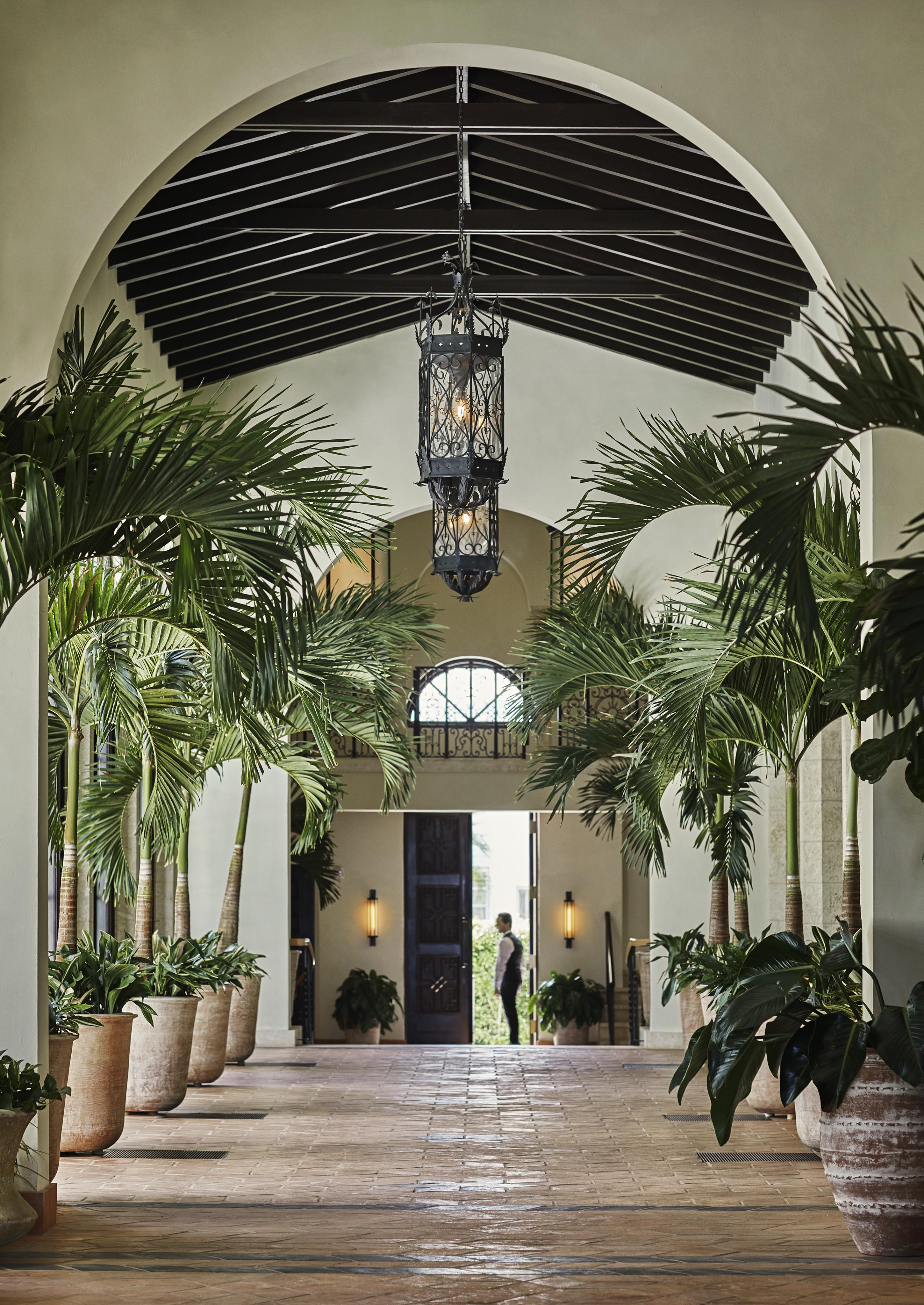 A Four Seasons Surf Club Miami corridor with potted palm trees and a hanging lantern. Hospitality staff stands at the end near a large door, with sunlight streaming in.