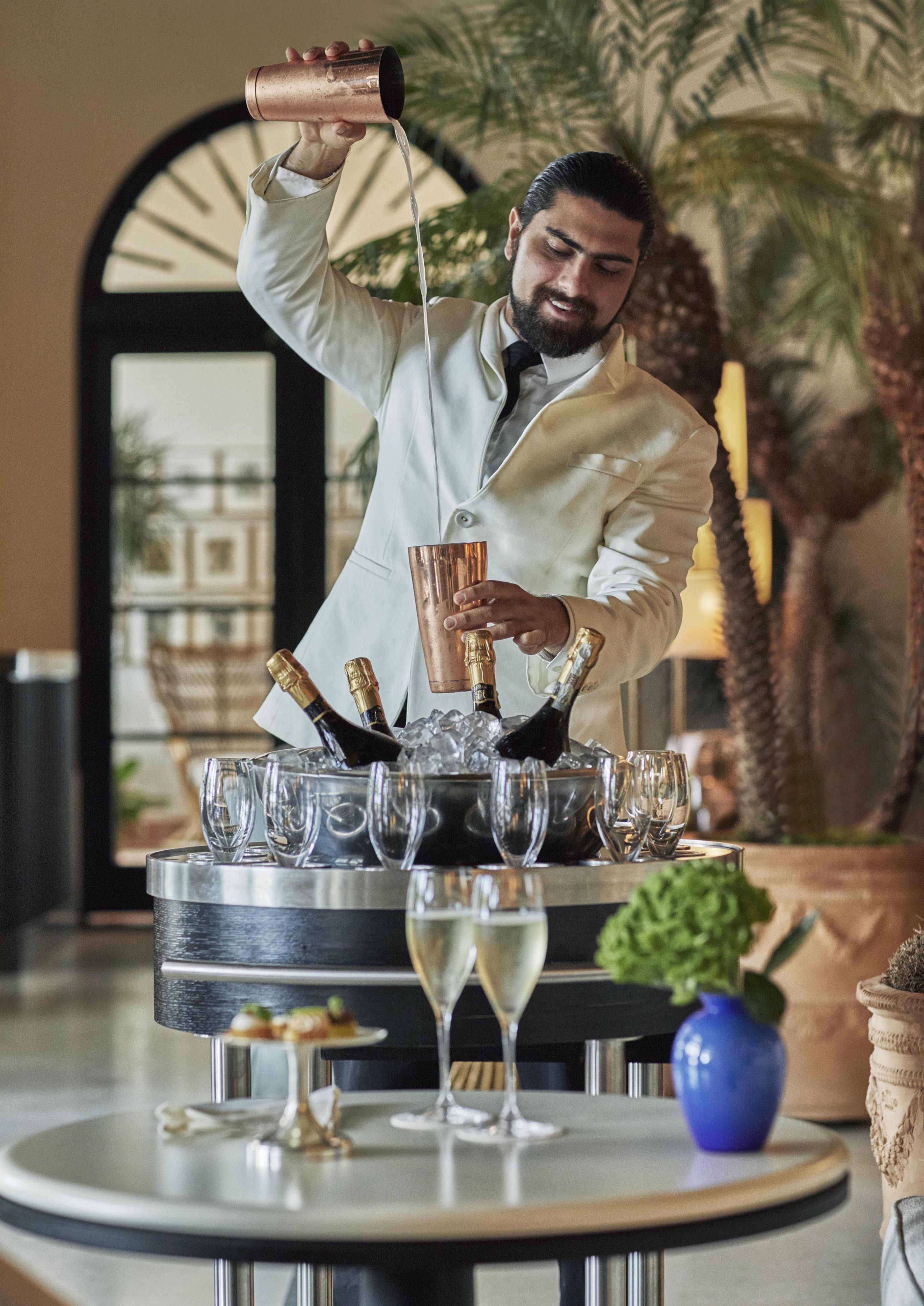 Four Seasons Miami resort bartender in white suit skillfully pours cocktail from shaker, with champagne bottles on ice nearby. Two filled glasses and small canapés on a table in a stylish lounge setting.