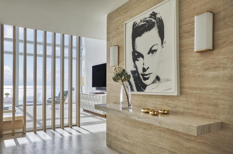 Modern interior with beige walls and natural light. Features a large black-and-white portrait, a sleek floating shelf with decor items, and a view of the sea through vertical wood slats.