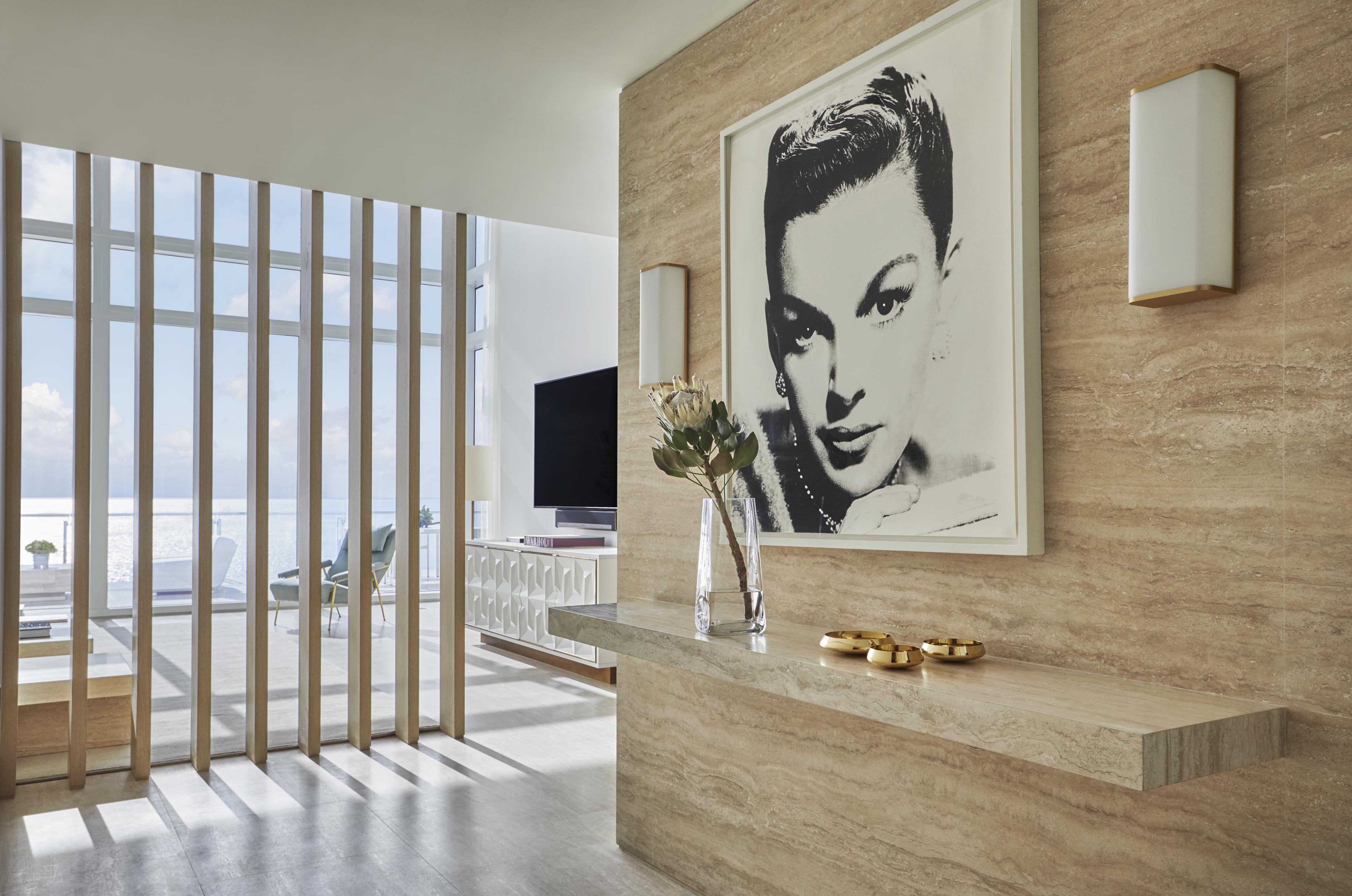 Modern interior with beige walls and natural light. Features a large black-and-white portrait, a sleek floating shelf with decor items, and a view of the sea through vertical wood slats.