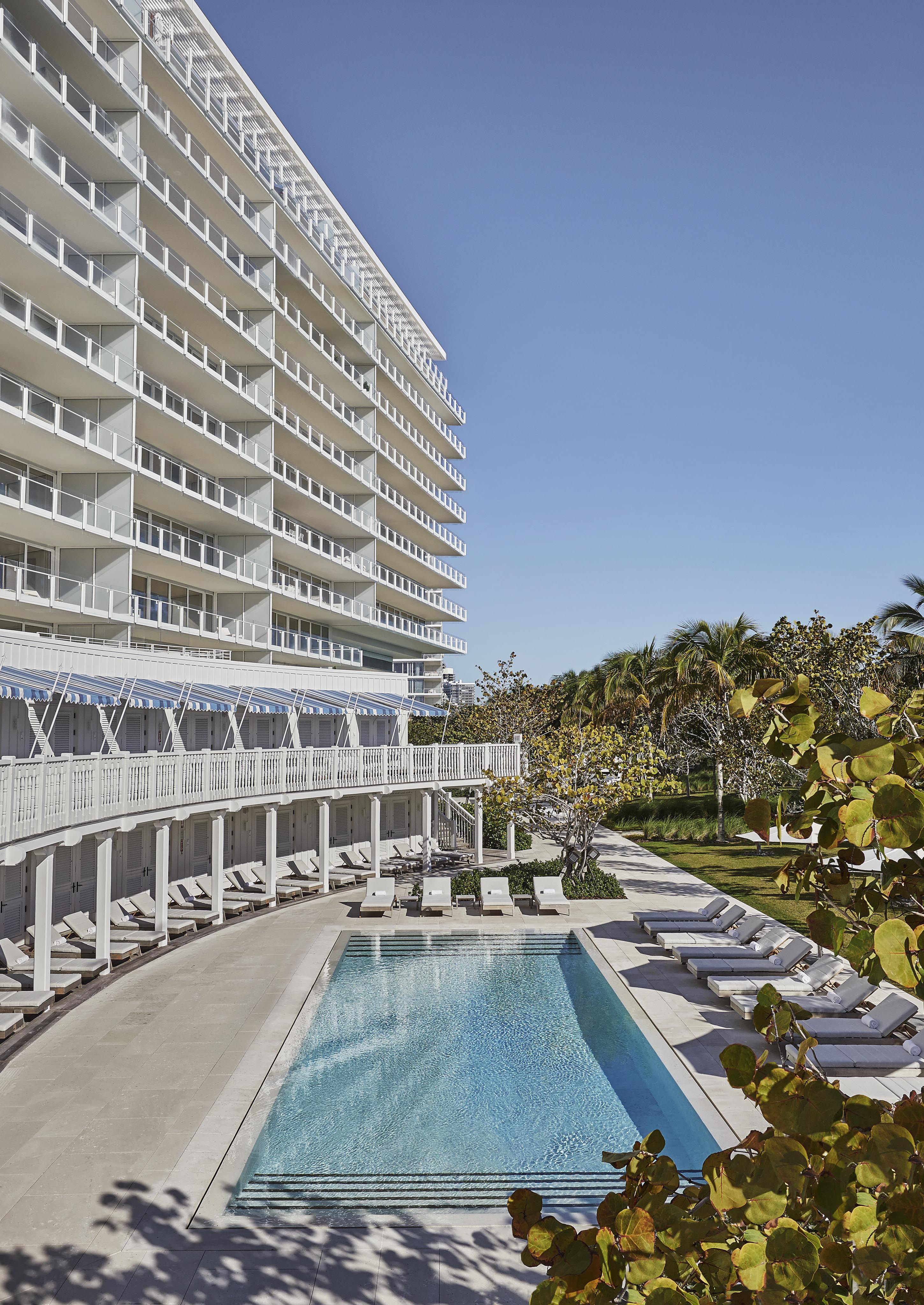 A modern hotel with multiple balconies overlooks a rectangular outdoor pool. Sun loungers are arranged around the pool, surrounded by trees and clear blue skies.